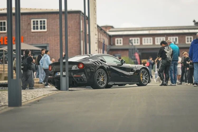 Schwarzer Sportwagen, wahrscheinlich Ferrari, parkt auf der Straße während eines Treffens, umgeben von mehreren Menschen.