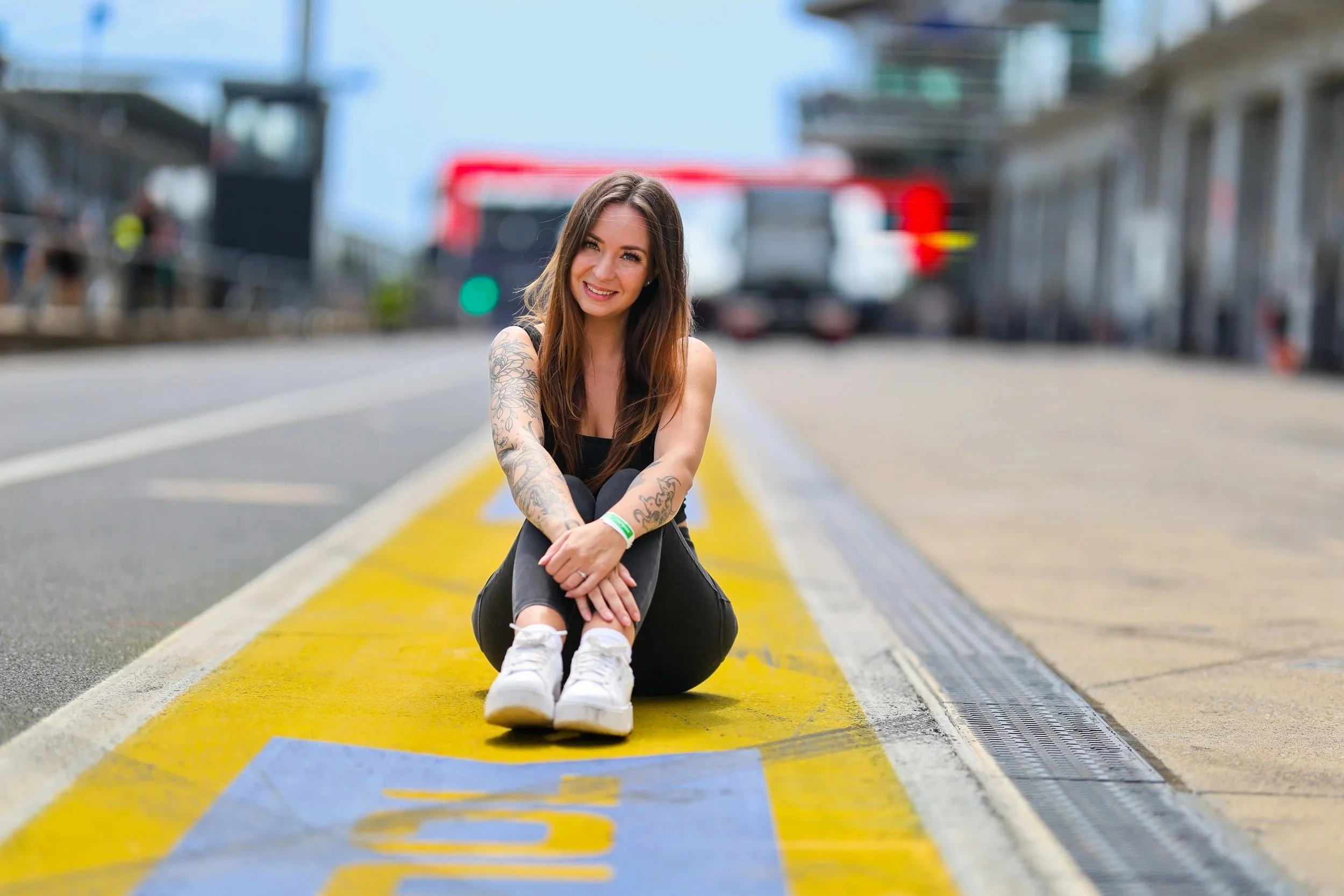 Junge Frau mit Tattoos sitzt lachend auf einer gelben Markierung auf der Straße, im Hintergrund sind Gebäude und ein Bus sichtbar.