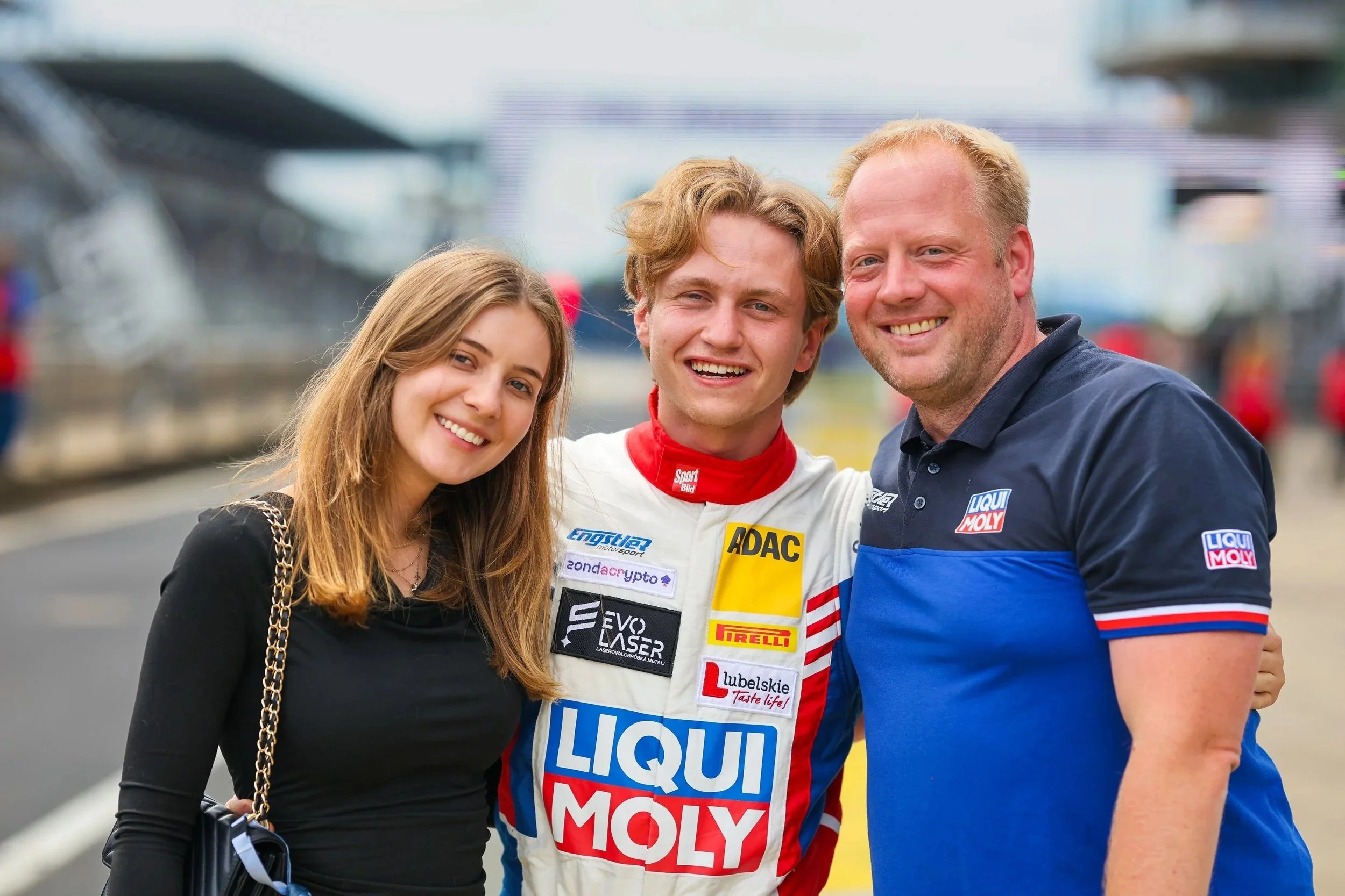 Drei Menschen, ein junger Mann im Rennfahreranzug und zwei Erwachsene, stehen zusammen bei einem Motorsport-Event und lächeln in die Kamera.
