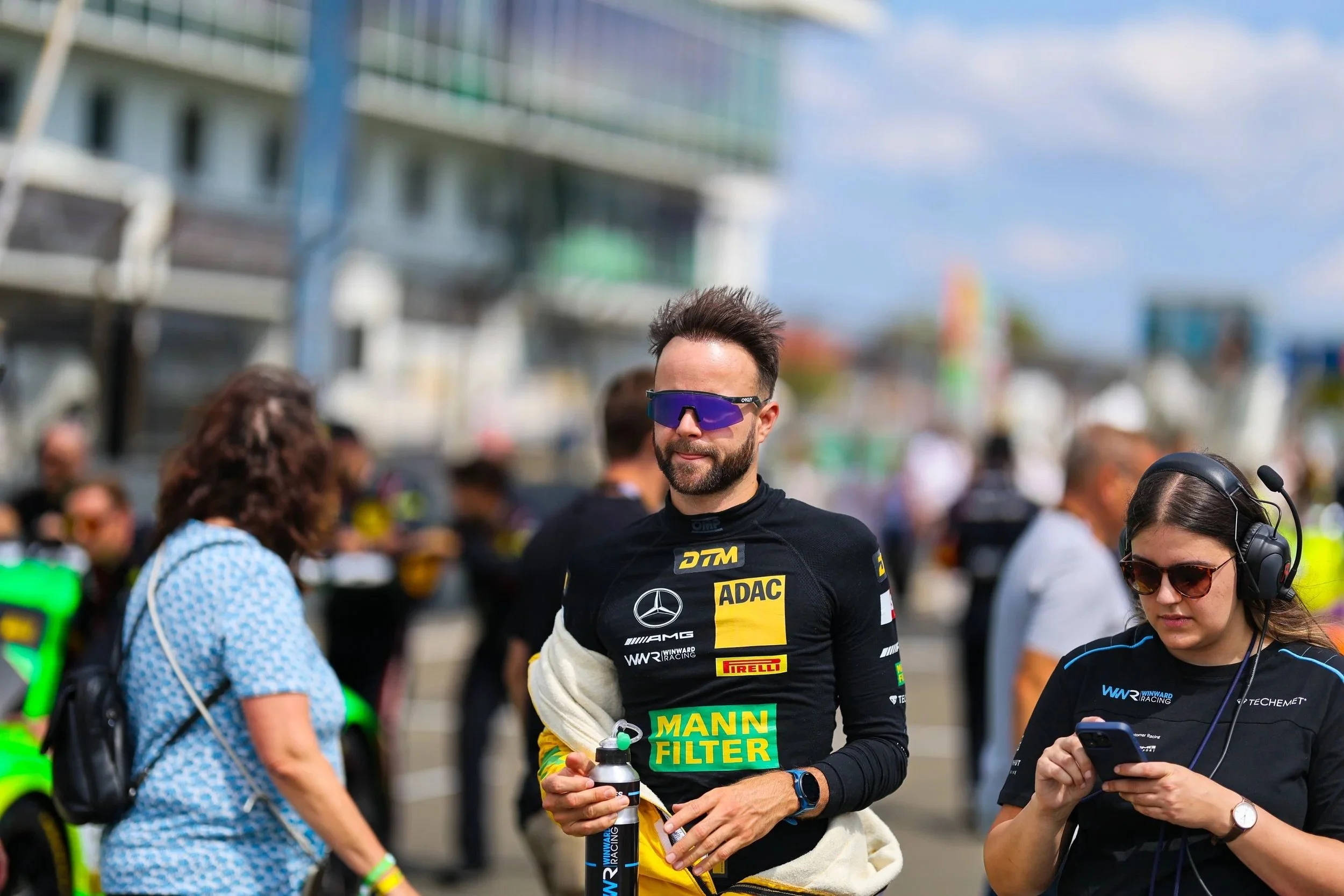 Motorsportfahrer in schwarzem Rennanzug mit Logos, trägt dunkle Sonnenbrille, steht an Rennstrecke, umgeben von Fans und Crew, mit Autos im Hintergrund.