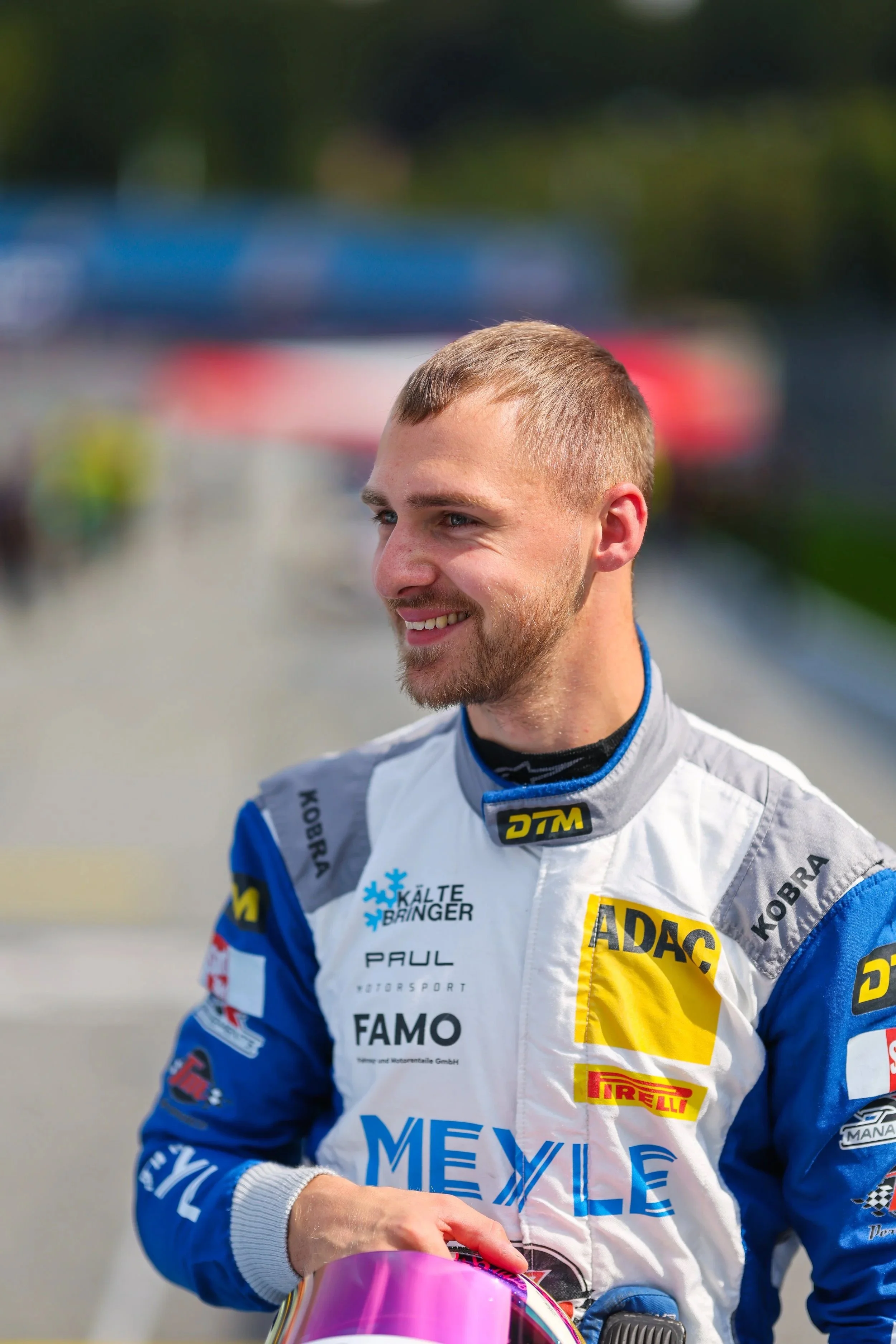 Ein lächelnder Autosportfahrer in Rennkleidung, hält einen pinken Rennhelm, im Hintergrund verschwommen. Es ist sonnig, vermutlich bei einem Rennen oder einer Veranstaltung.