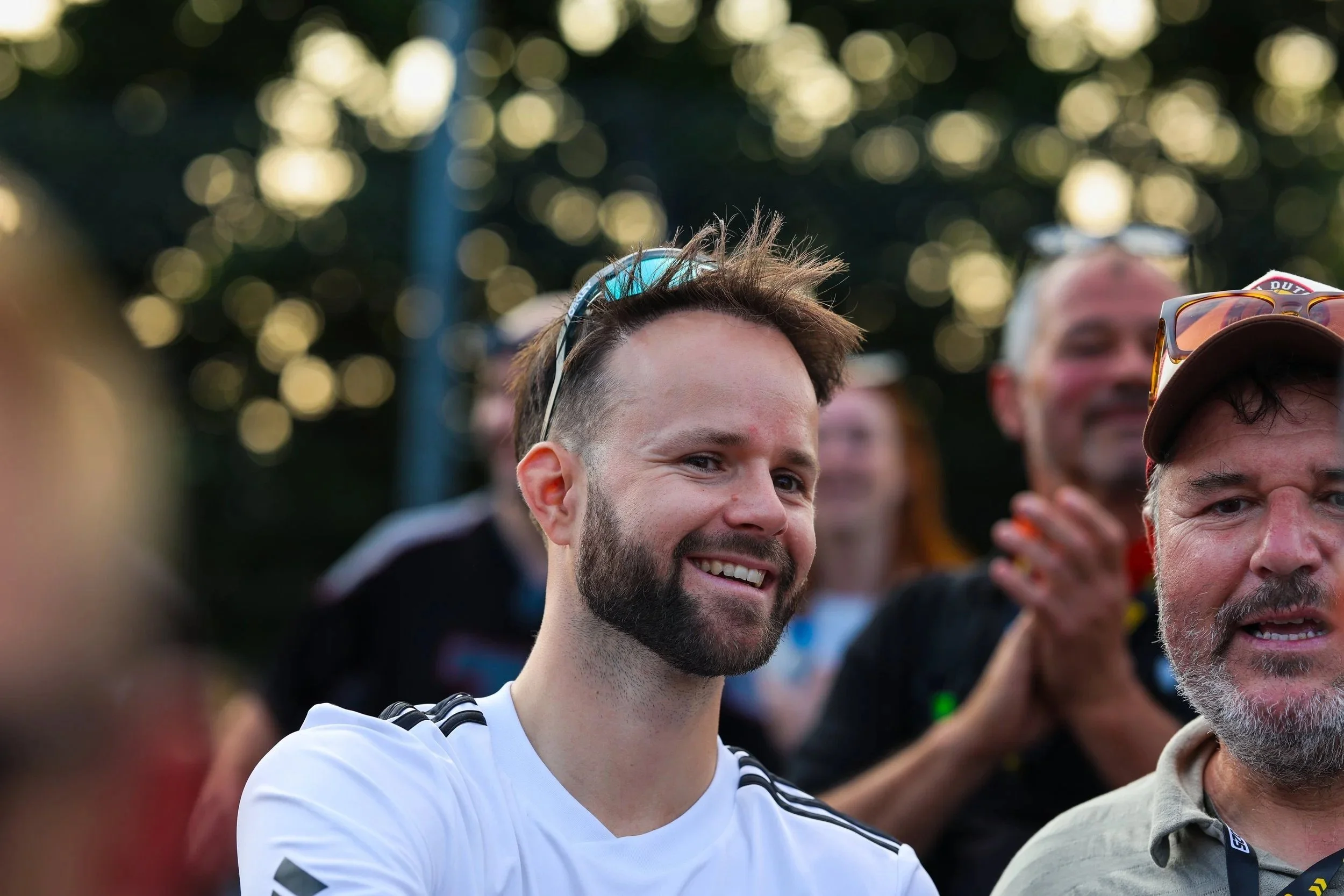 Gruppe von Menschen beim Outdoor-Event, im Mittelpunkt ein lachender junger Mann mit Bart, Sonnenbrille auf dem Kopf, trägt ein weißes T-Shirt, im Hintergrund Sonnenlicht durch Bäume.