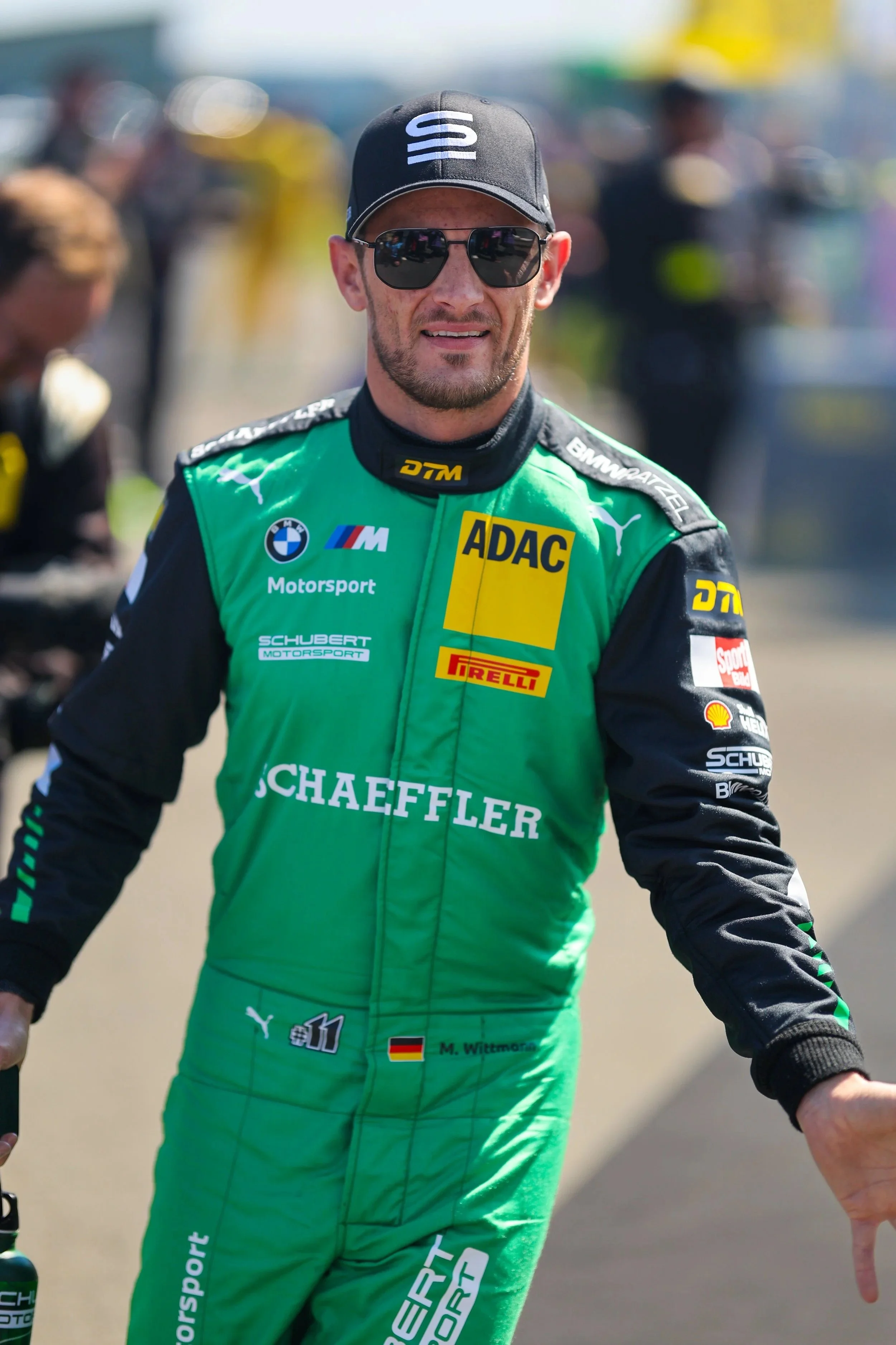 Motorsportfahrer in grüner Rennanzug, Sonnenbrille, schwarzer Cap, im Fahrerlager, mit Sponsoren und Teamlogos.