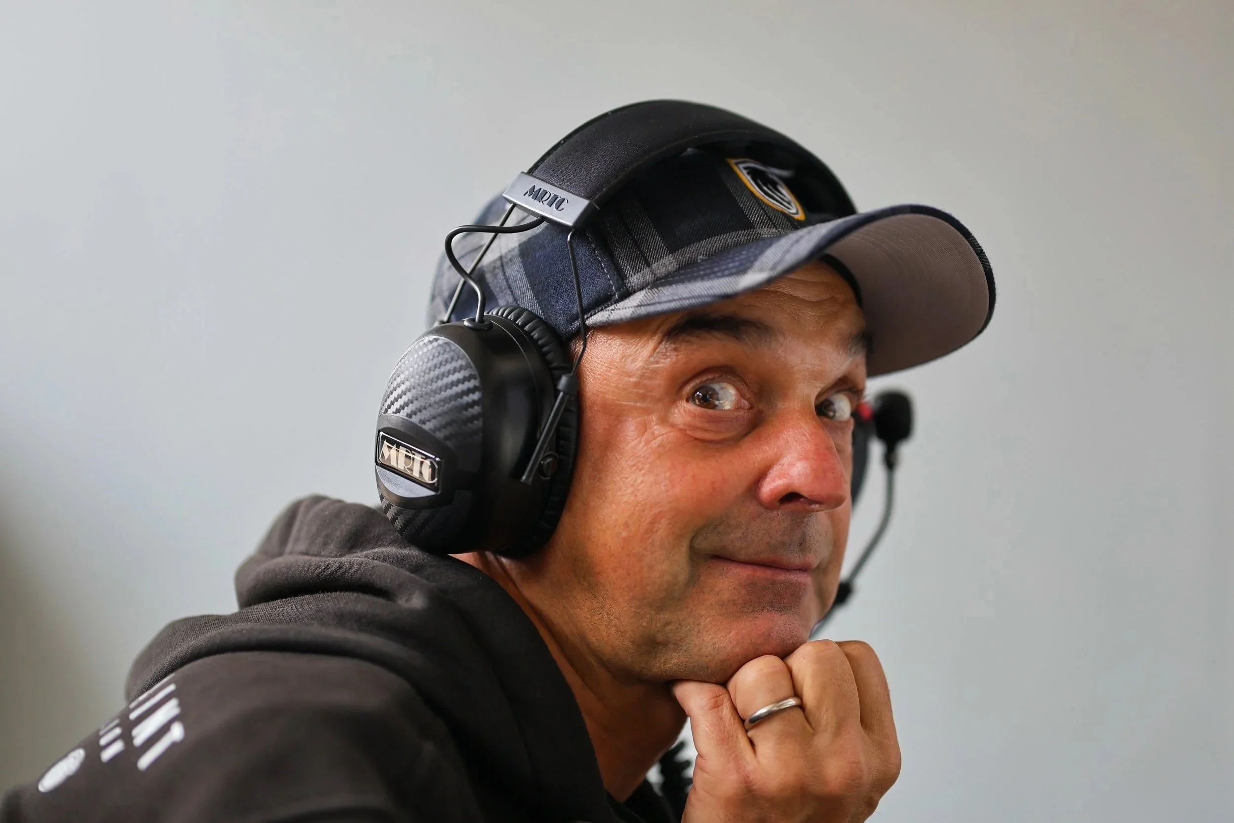 Mann trägt Kopfhörer, eine Baseballkappe, trägt ein schwarzes Hoodie und blickt in die Kamera mit einem schmunzelnden Gesicht, die Hand ruht unter dem Kinn.