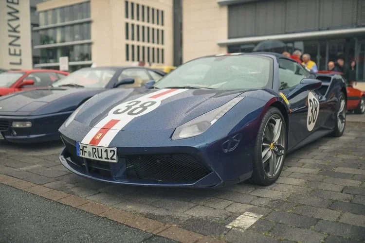 Ein blauer Sportwagen, wahrscheinlich ein Lamborghini, parkt auf einem Platz vor einem modernen Gebäude, umrahmt von anderen Autos. Das Fahrzeug trägt Motorsport-Nummern und eine charakteristische Rennlackierung.