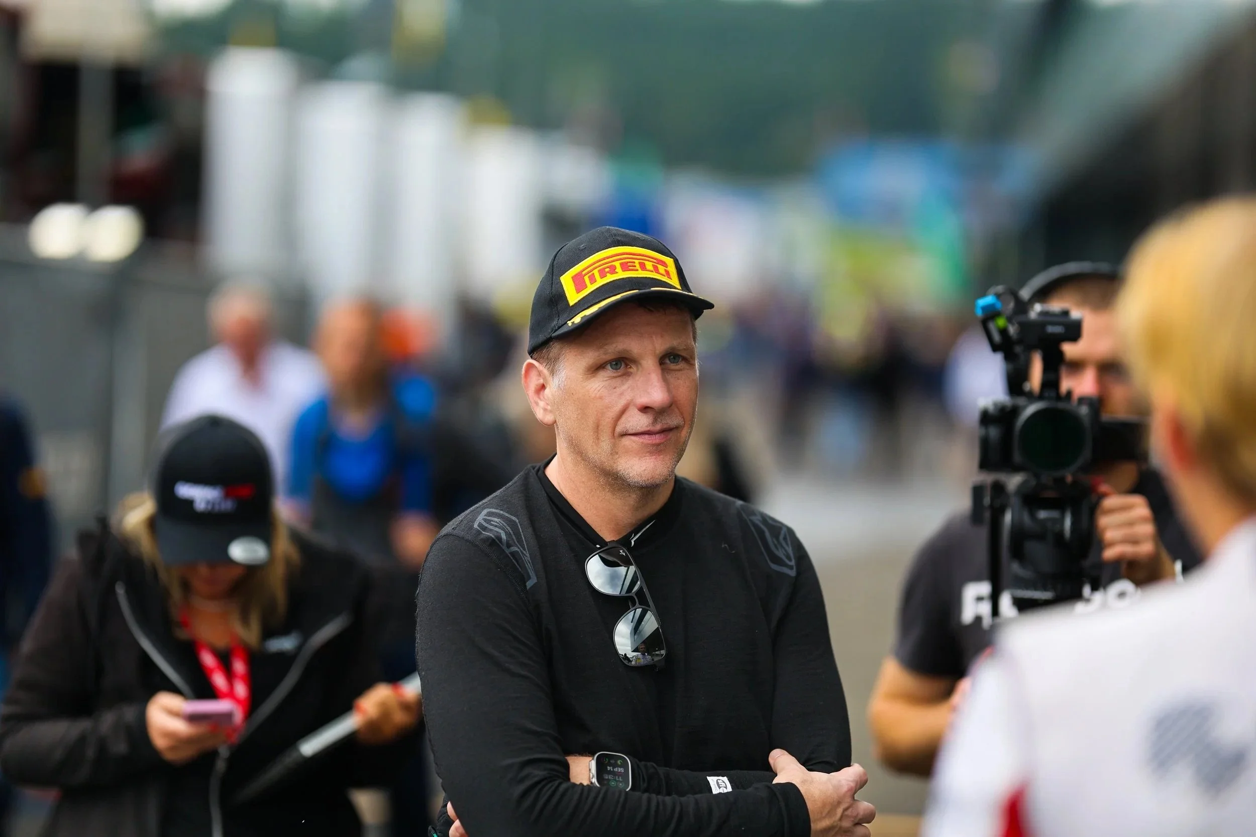Ein Mann steht bei einem Motorsport-Event, trägt einen black T-Shirt mit reflektierenden Sonnenbrillen, eine schwarze Mütze mit Pirelli-Logo und hat eine Uhr am Handgelenk, während er im Gespräch mit einer Person ist, die eine Kamera trägt.