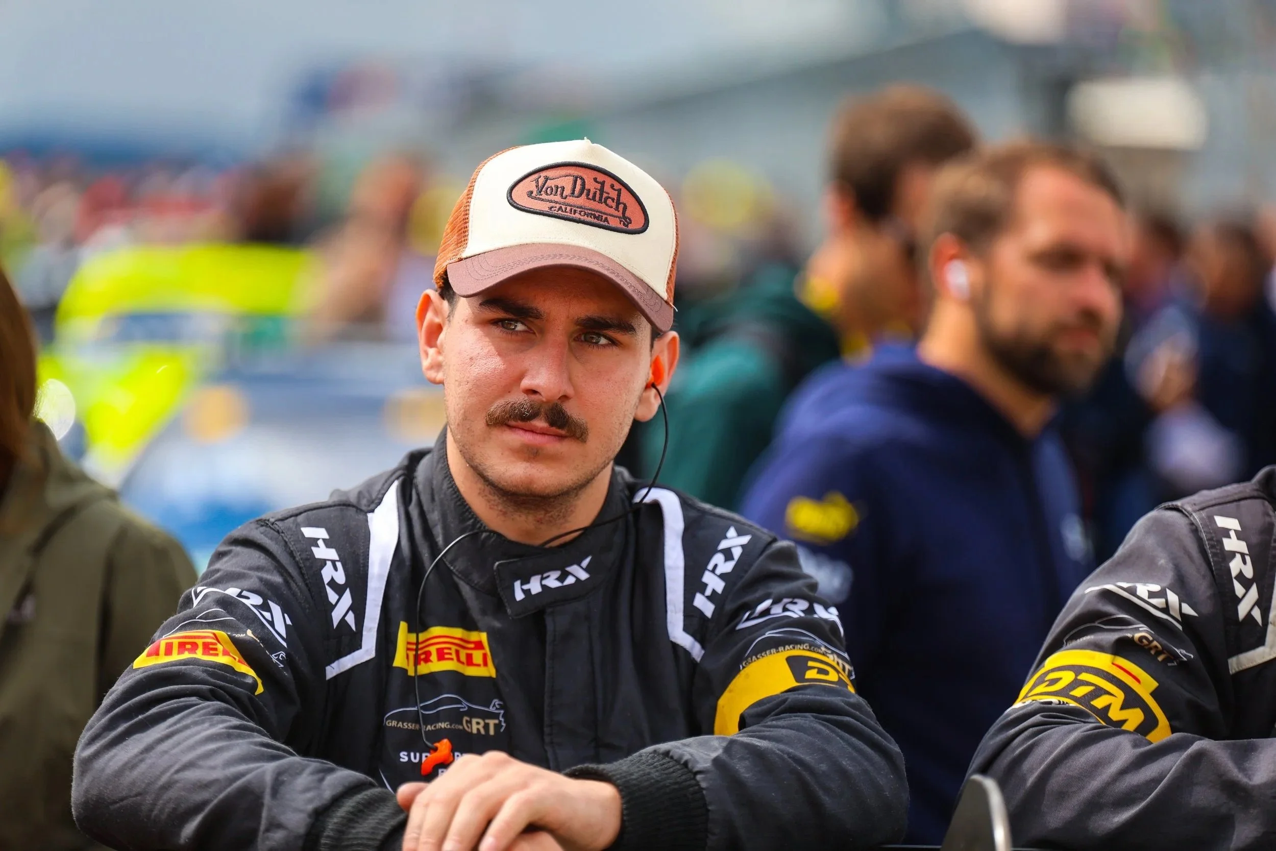 Ein junge Mann in einem schwarzen Rennfahreranzug mit Pirelli-Logo sitzt bei einem Motorsport-Event, trägt einen weißen und orangefarbenen Cap, hat einen Schnurrbart und hört mit einem Ohrhörer Musik.