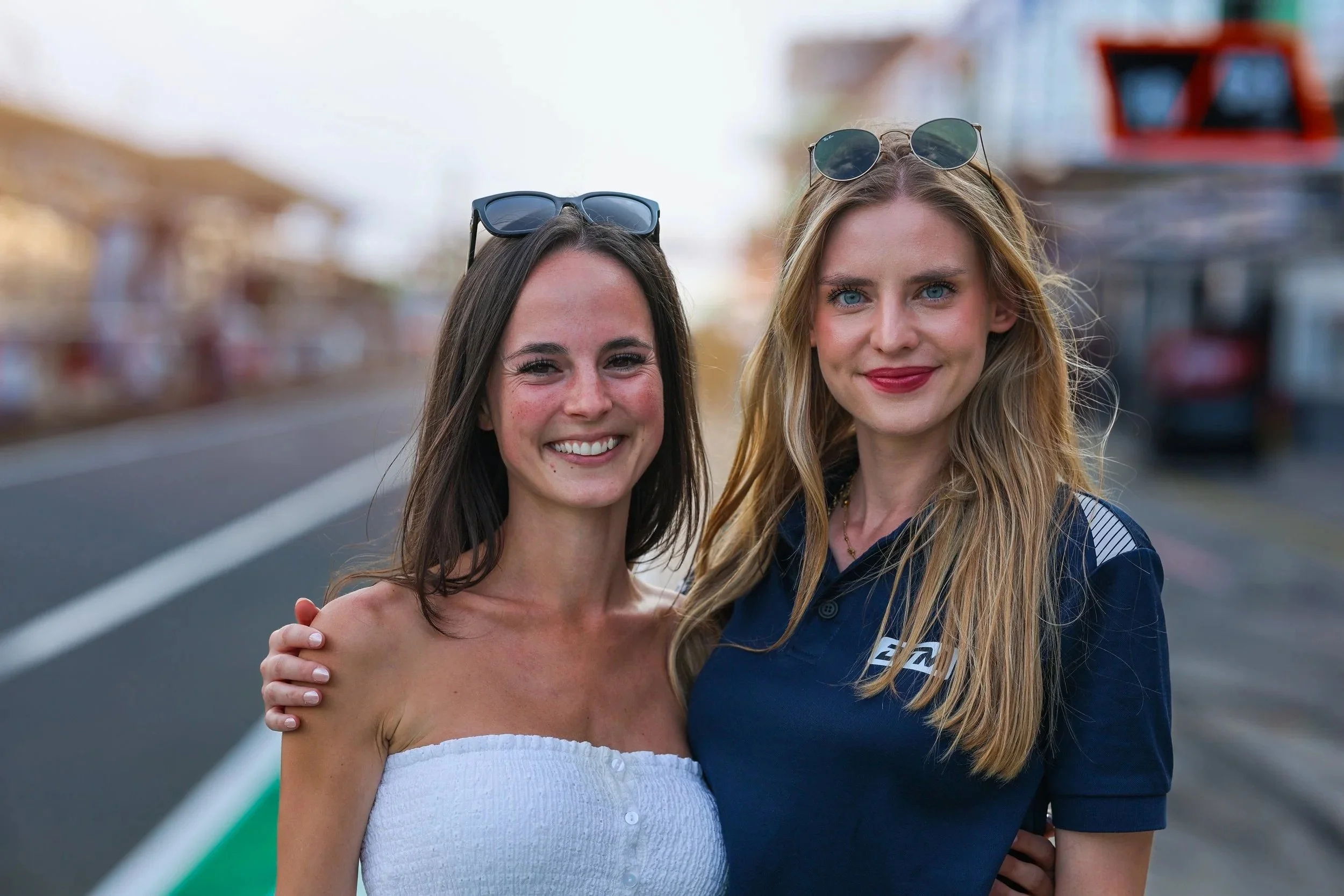Zwei lachende Frauen stehen eng nebeneinander auf einer Straße, eine trägt ein weißes Strandkleid, die andere ein dunkelblaues Polo-Shirt, beide tragen Sonnenbrillen auf dem Kopf, im Hintergrund sind Gebäude und ein Himmel sichtbar.