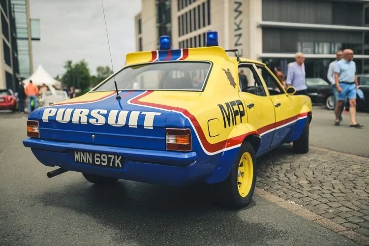 Ein gelb-blaues Auto mit Polizei-Design, das auf einem Gehweg parkt, mit Menschen im Hintergrund.