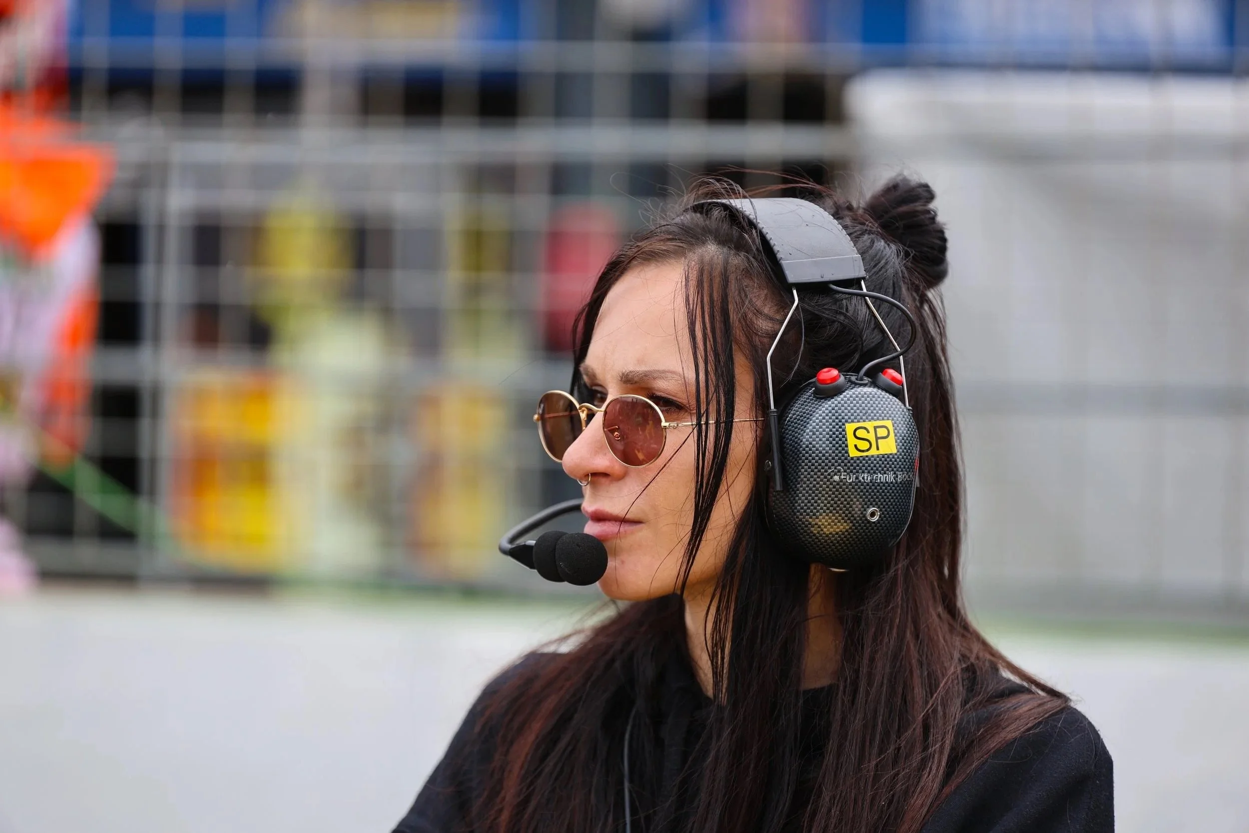 Junge Frau mit dunkelhaar, Sonnenbrille, und Headset an einem Rennstrecke, hinter Gitterzaun.
