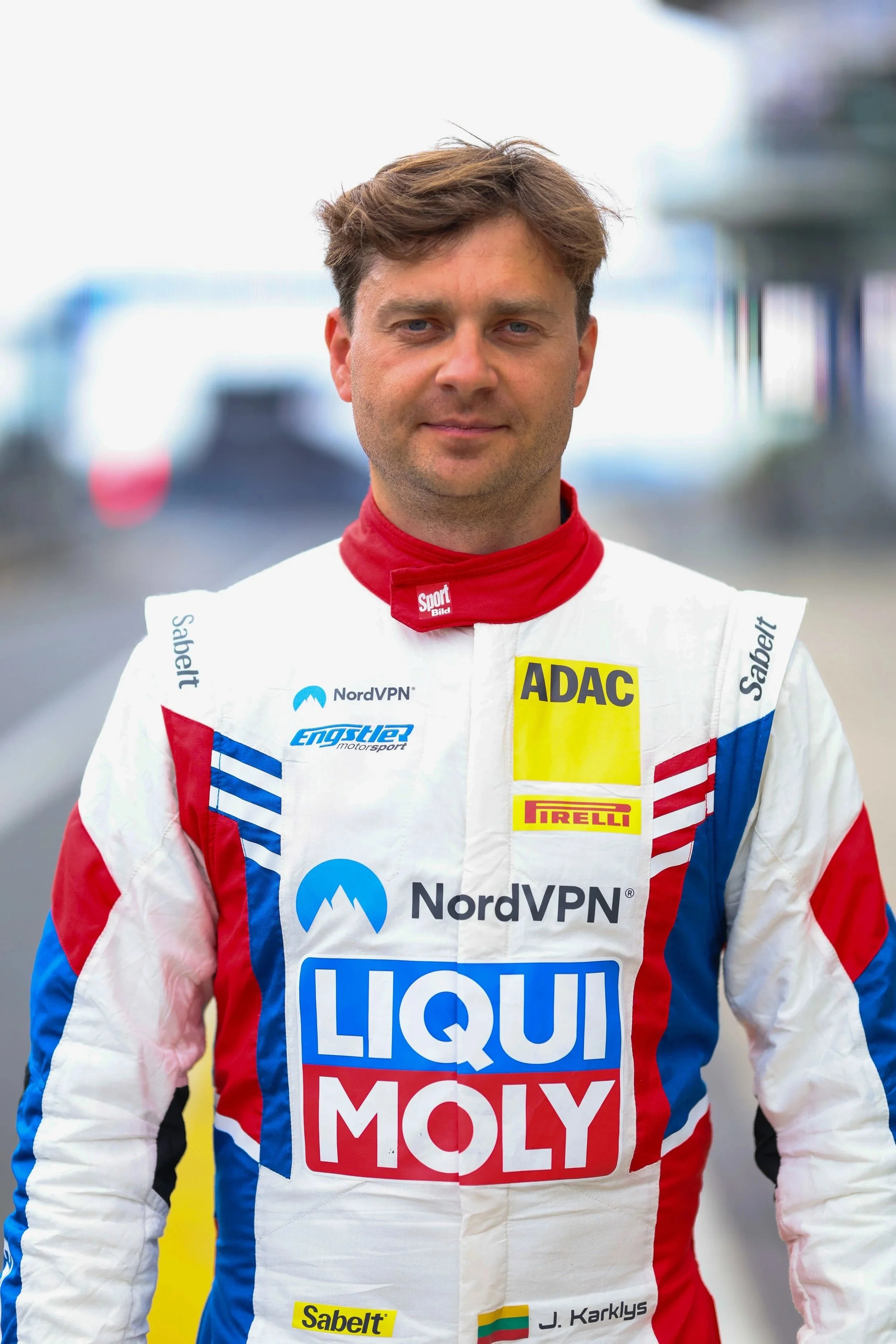 Ein Rennfahrer steht in Rennbekleidung mit Sponsorenlogos, darunter Liqui Moly, ADAC und Pirelli, in einer Boxenstraße mit Autos im Hintergrund.