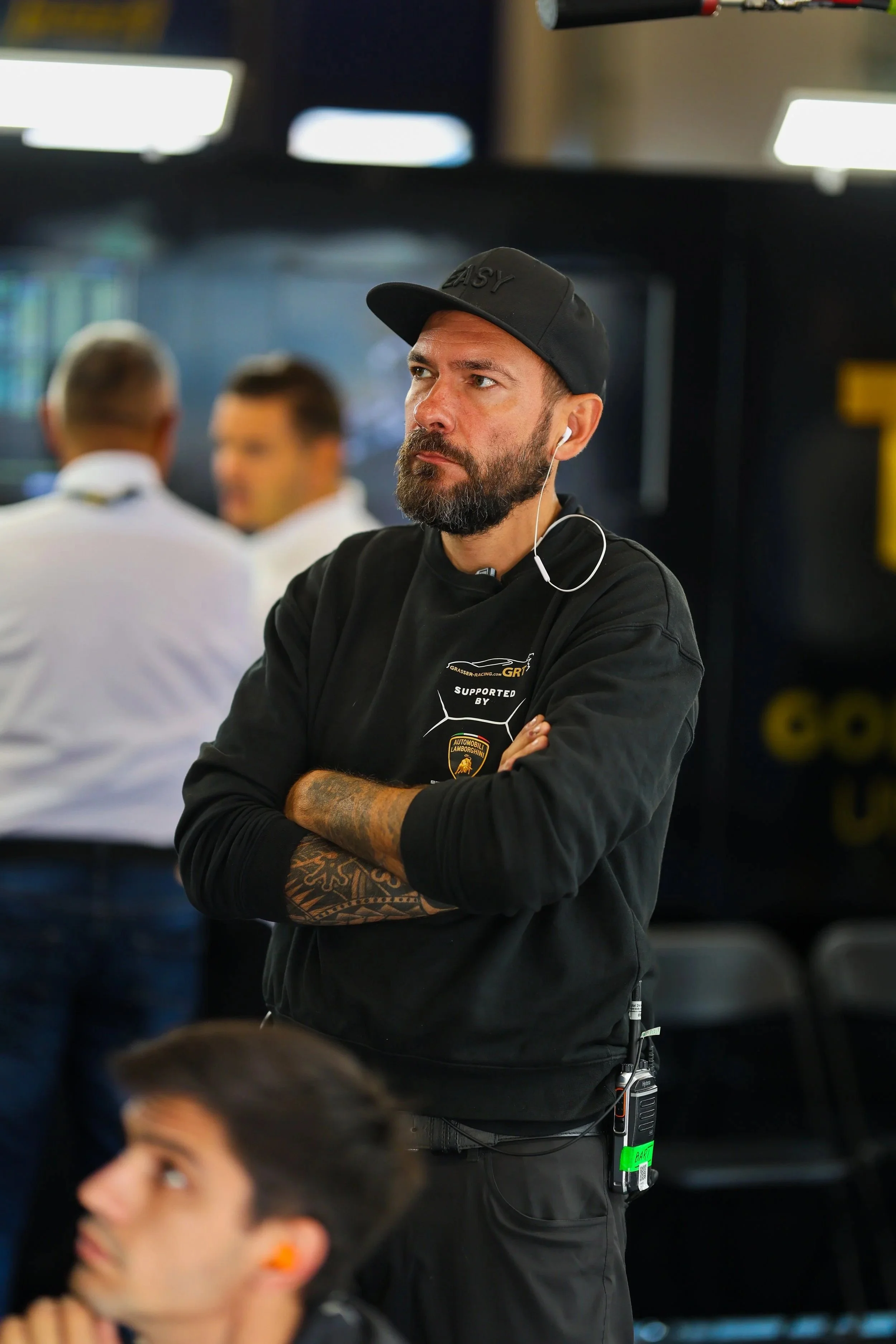 Mann mit dunklem Cap, schwarzen Pullover und Tattoo am Arm, der in einem Motorsport-Event steht, mit anderen im Hintergrund.