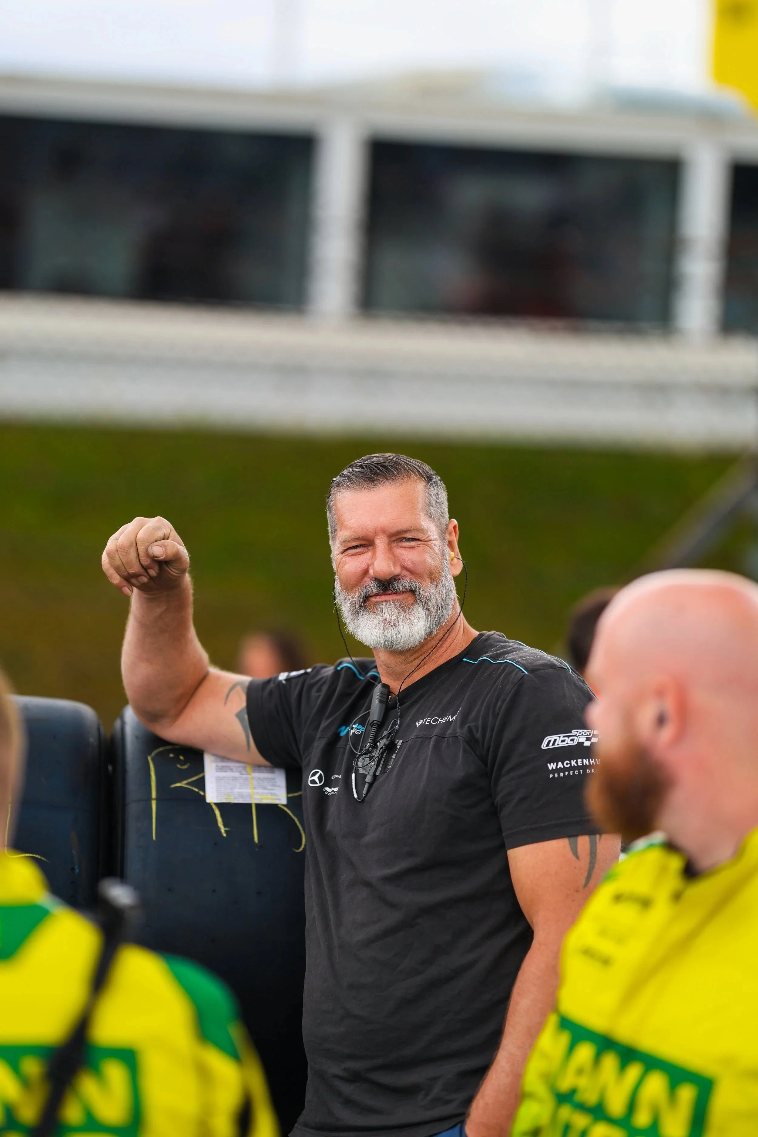 Mann mit grauem Bart und schwarzem T-Shirt, der einen Arm muskulös zeigt, während er lächelt, auf einem Rennen oder Event, mit anderen Menschen in leuchtend gelben Shirts im Vordergrund.