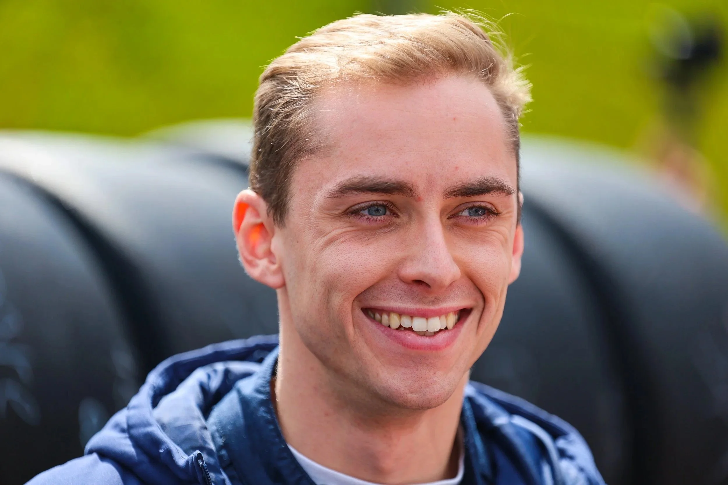 Lächelnder junger Mann mit blonden Haaren und blauen Augen, trägt eine blaue Jacke, im Hintergrund ein dunkler Reifen