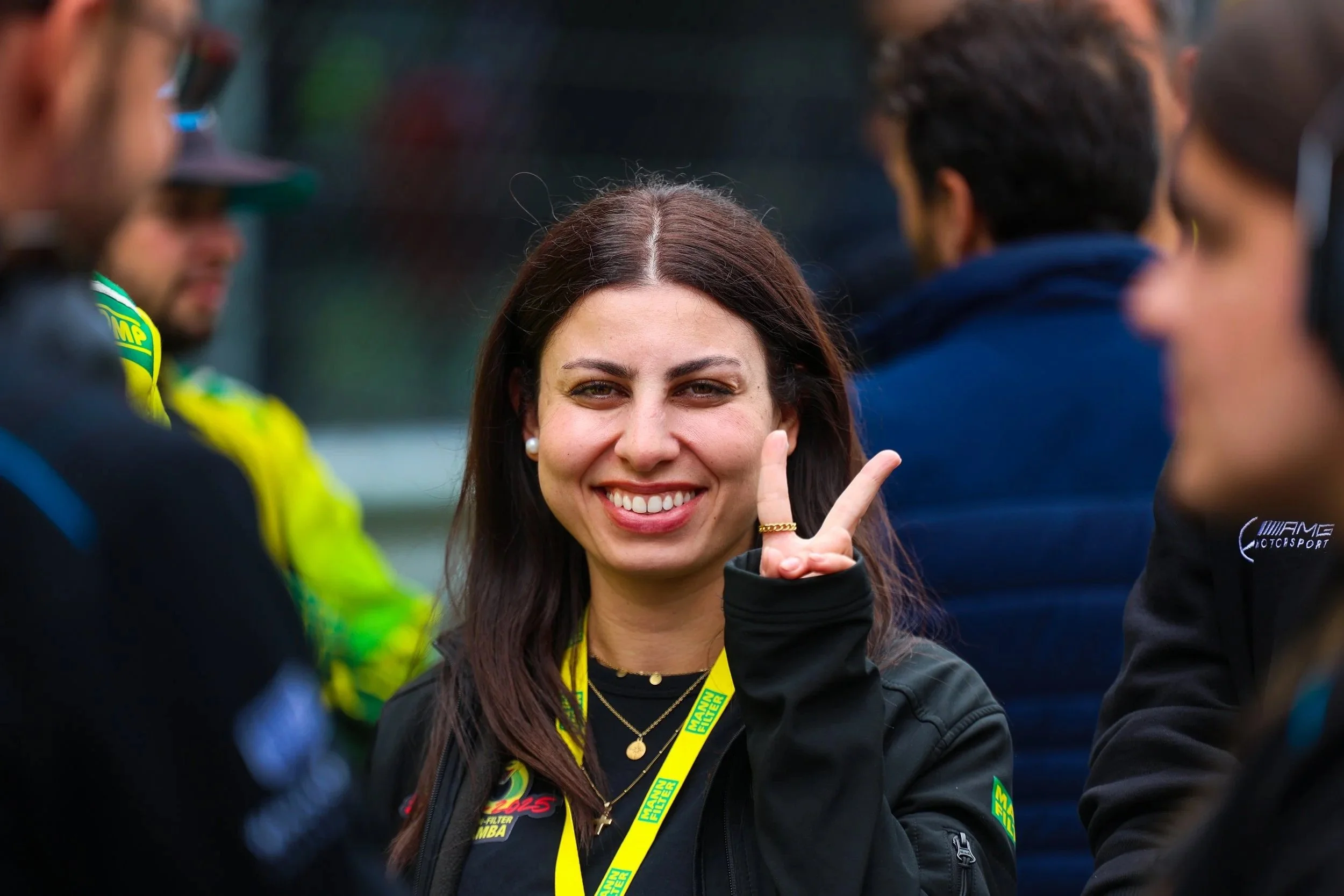 Eine lächelnde Frau macht ein Peace-Zeichen mit ihrer Hand in einer Gruppe von Menschen. Sie trägt eine schwarze Jacke und hat eine gelbe Lanyard um den Hals, vermutlich bei einer Motorsportveranstaltung.