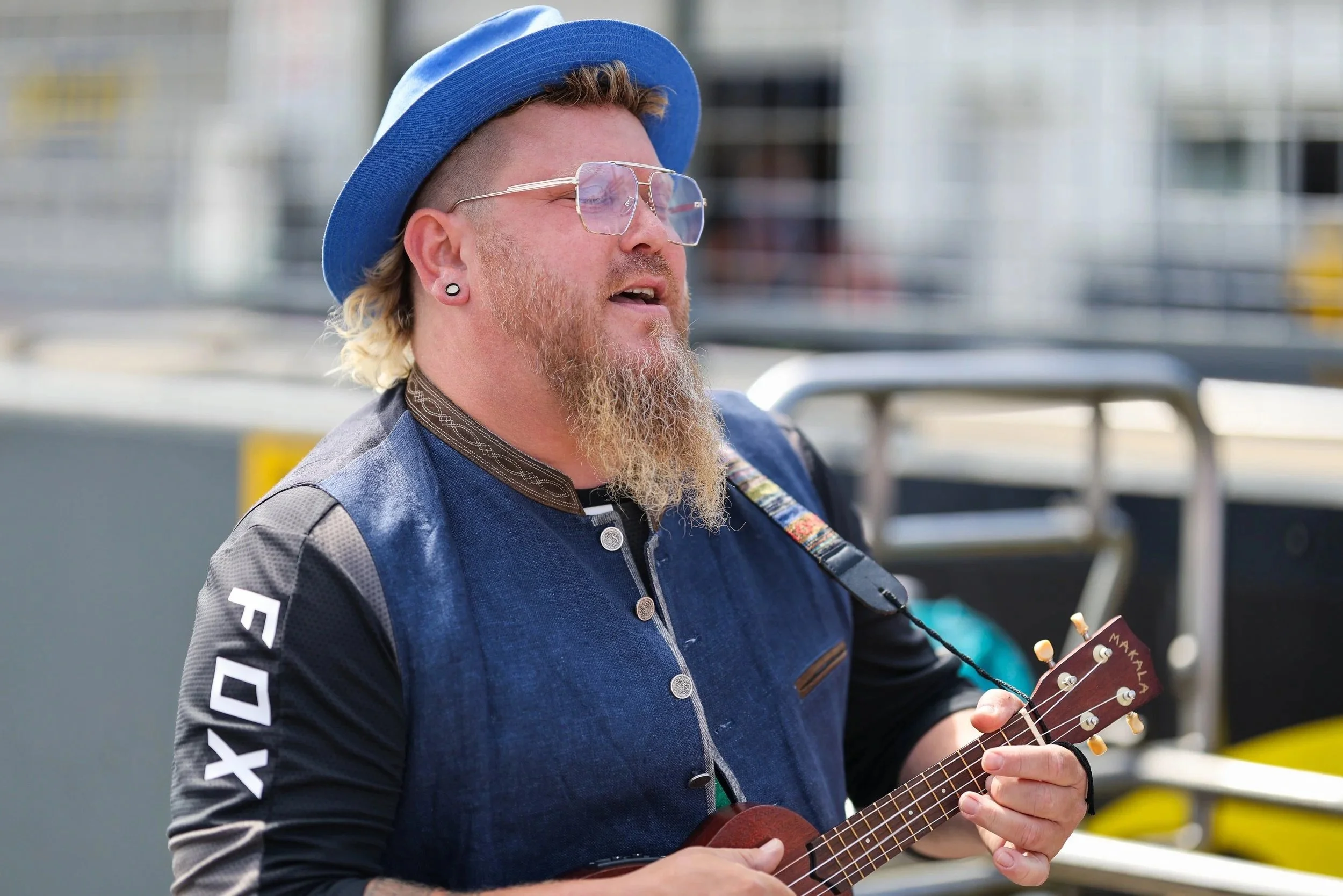 Ein Mann mit Brille, blonden lockigen Haaren, Vollbart und Ohrring, spielt auf einer Ukulele.