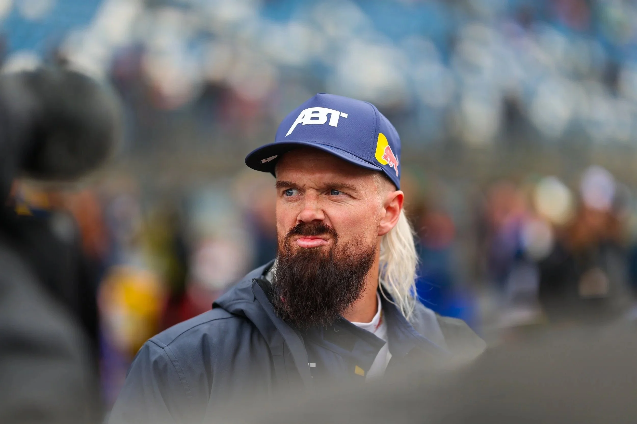 Ein Mann mit blondem Haar, Vollbart und blauen Augen, trägt einen blauen Cap mit den Logos von ABT und Red Bull, und eine schwarze Jacke. Im Hintergrund sind unscharfe Personen und Oberflächen.
