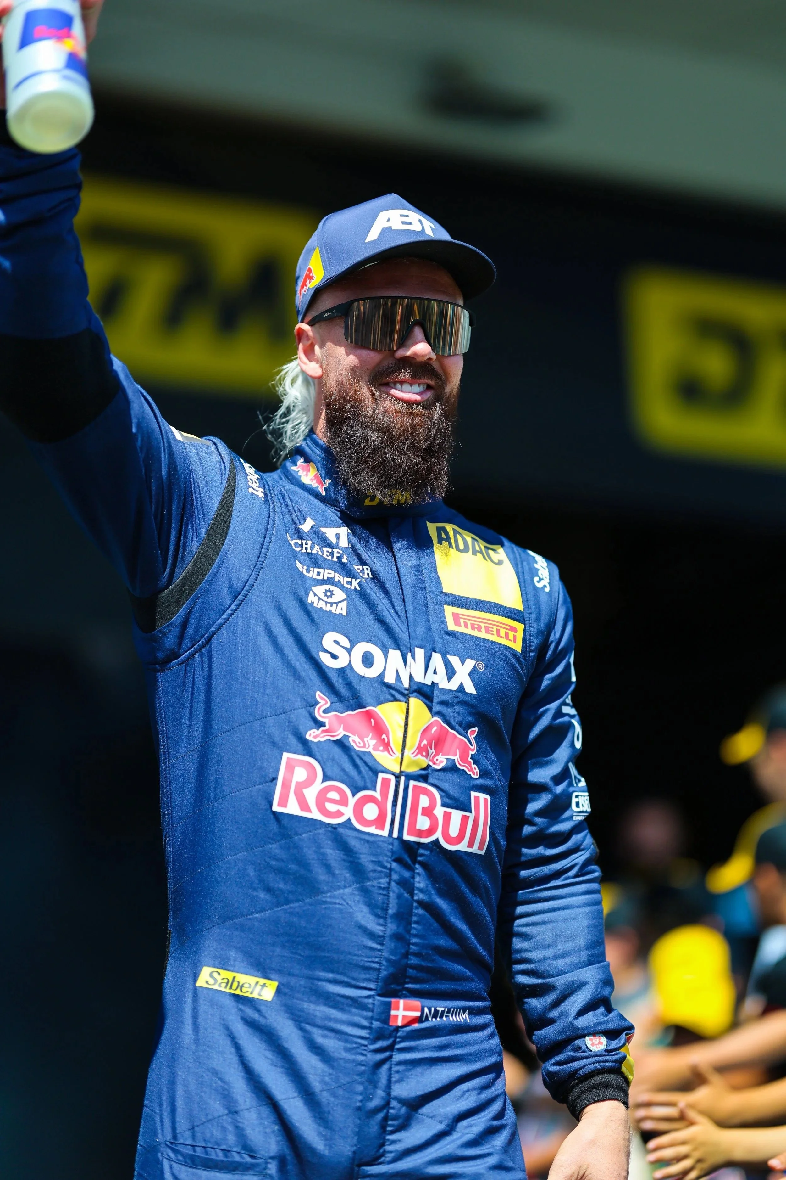 Motorsportfahrer in blauer Rennkutte mit Red Bull und anderen Logos, trägt Sonnenbrille, winkt, vor Zuschauer
