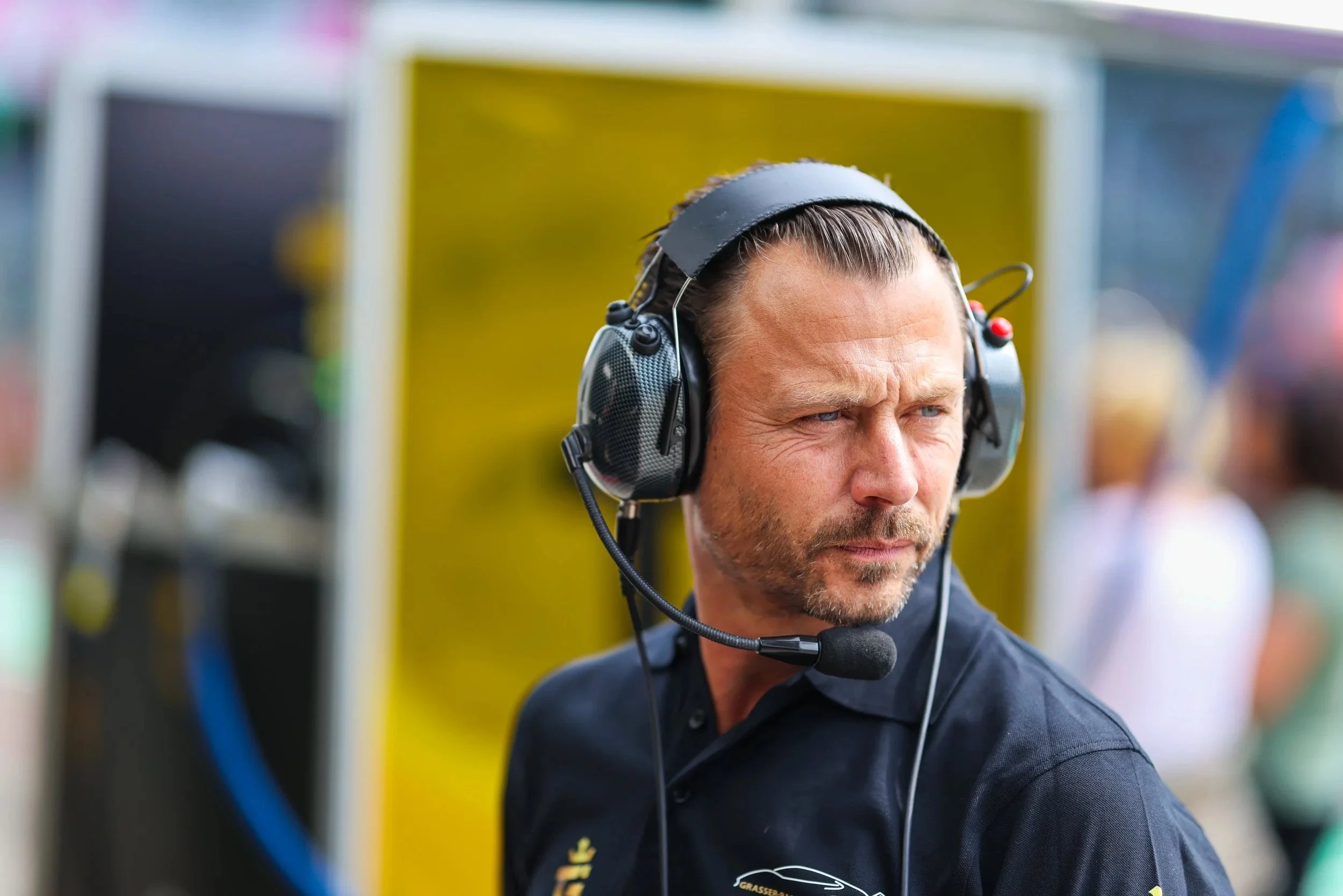 Ein Mann trägt ein Headset mit Mikrofon und schaut nach rechts, im Hintergrund sind Menschen und bunte Flächen sichtbar.