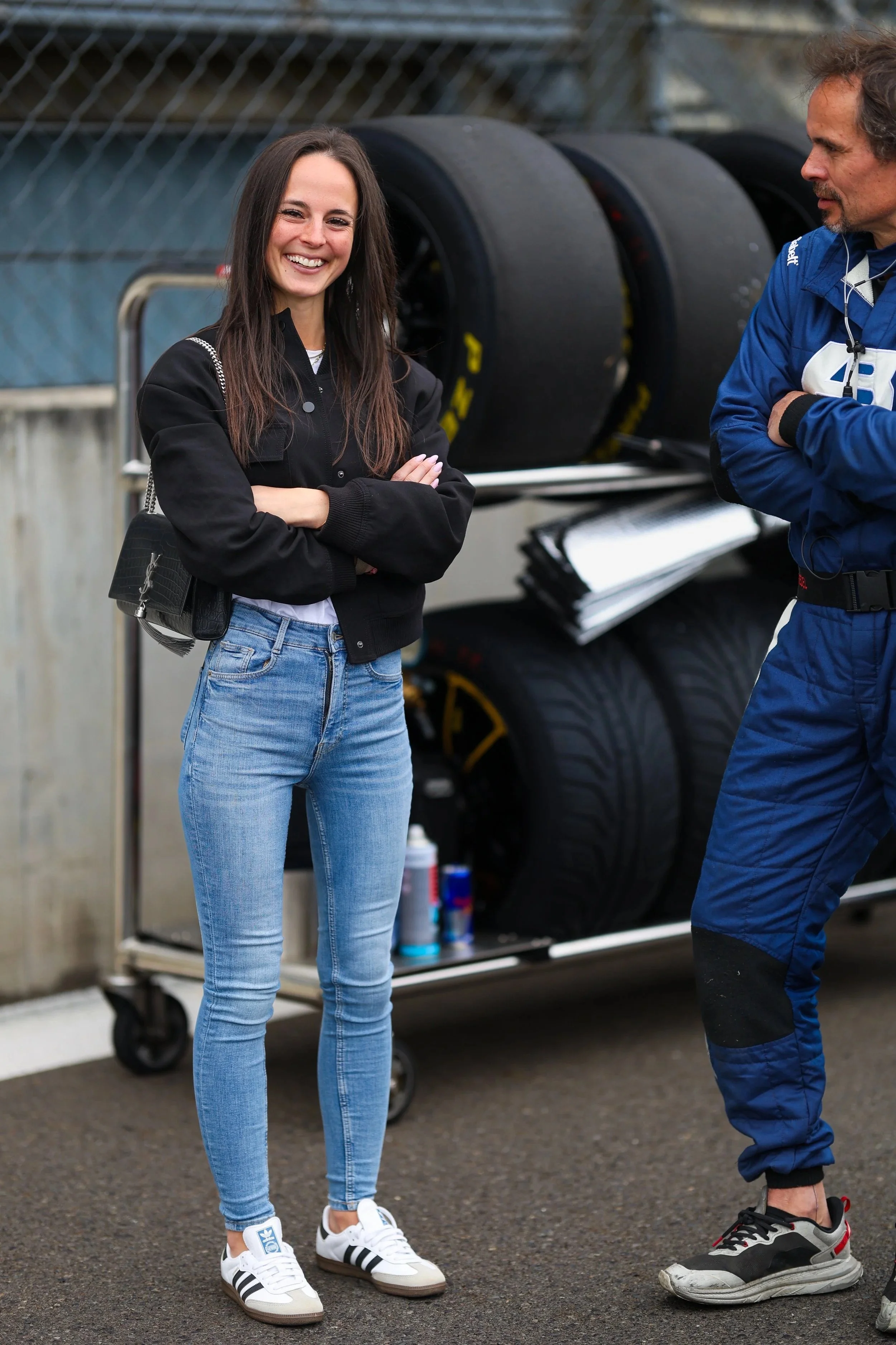 Junge Frau mit dunklen Haaren, Jeans, schwarzer Jacke, weißen Sneakers stand lächelnd mit verschränkten Armen neben einen Mann in blauer Rennfahrerbekleidung vor einem Reifenregal.