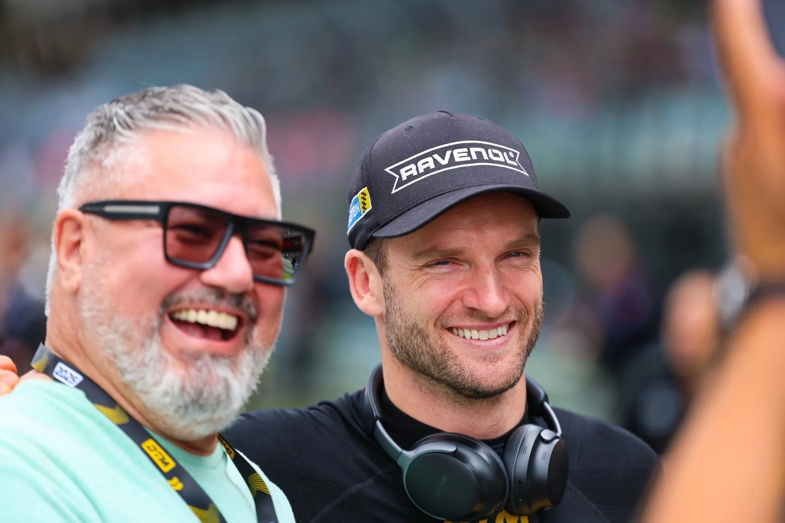 Zwei lachende Männer beim Fotoshooting, einer trägt Sonnenbrille, der andere eine Cap mit Aufdruck, beide haben Headsets um den Hals, draußen bei einem Event.