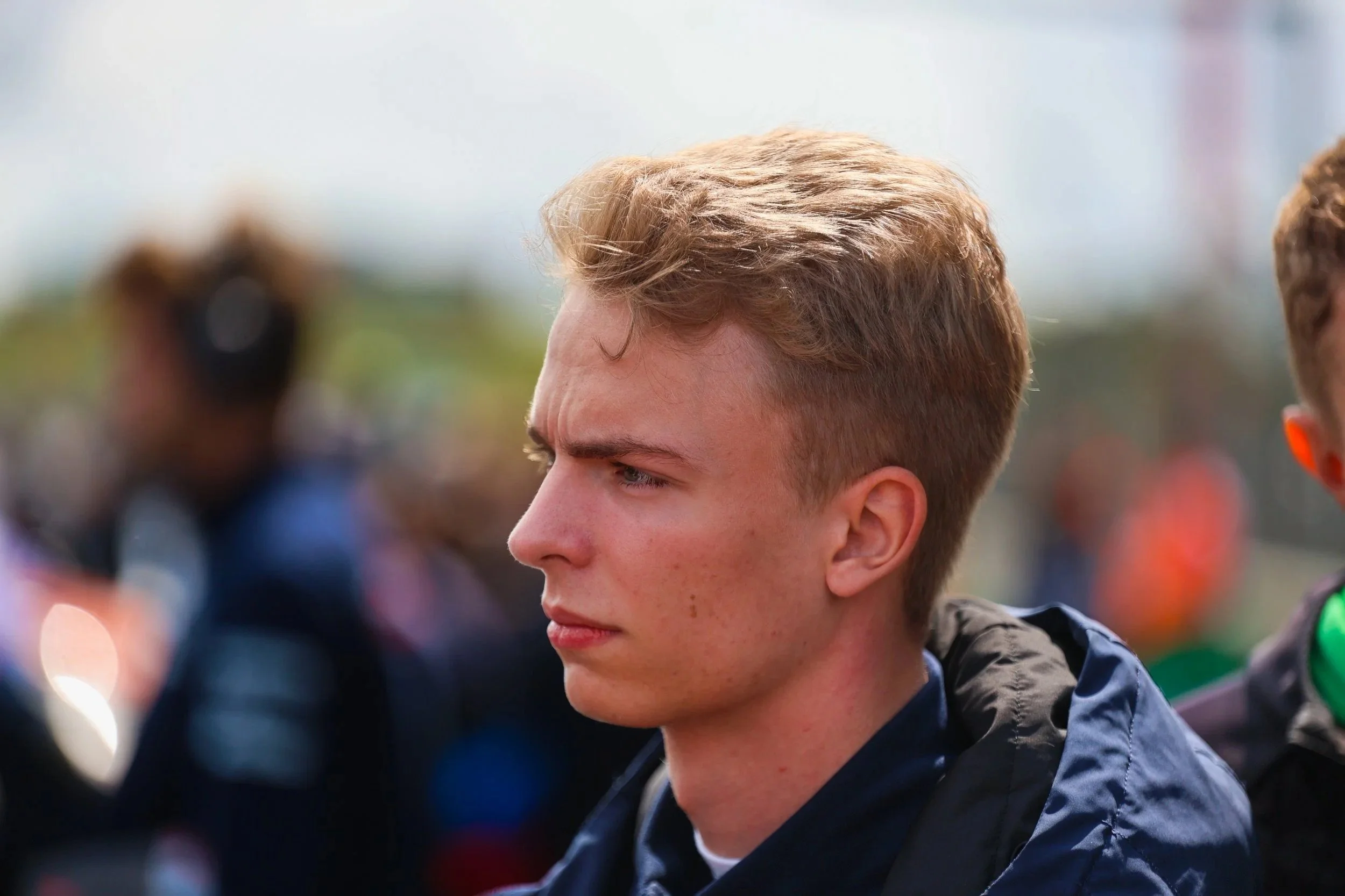 Nahaufnahme eines jungen Mannes mit kurzen, hellbraunen Haaren, der ernst oder konzentriert schaut, im Hintergrund andere Personen bei einem Outdoor-Event.