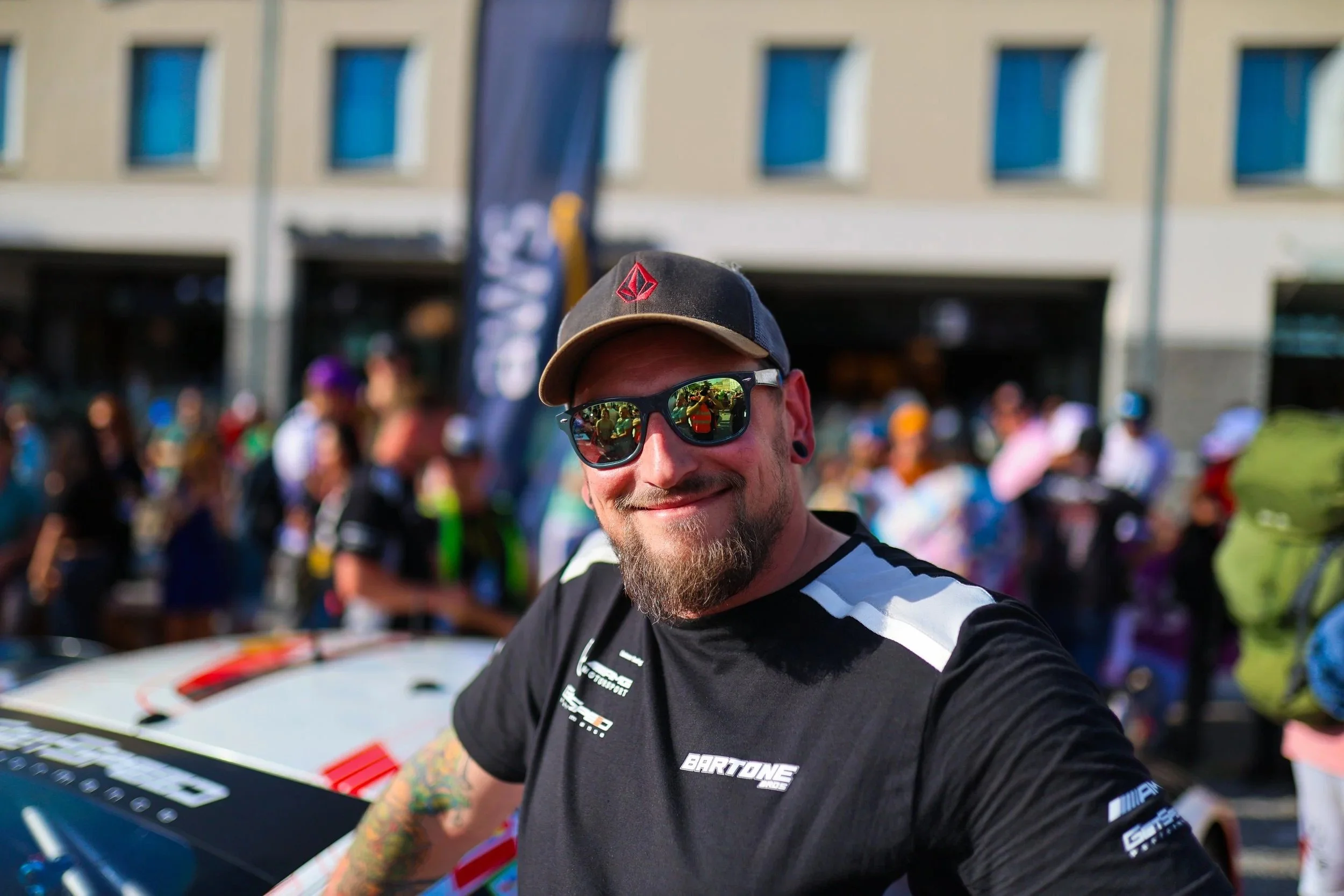 Lächelnder junger Mann mit Sonnenbrille, schwarzem Cap, schwarzen T-Shirt und Tattoos auf dem Arm, im Hintergrund eine Menschenmenge bei einem Outdoor-Event.