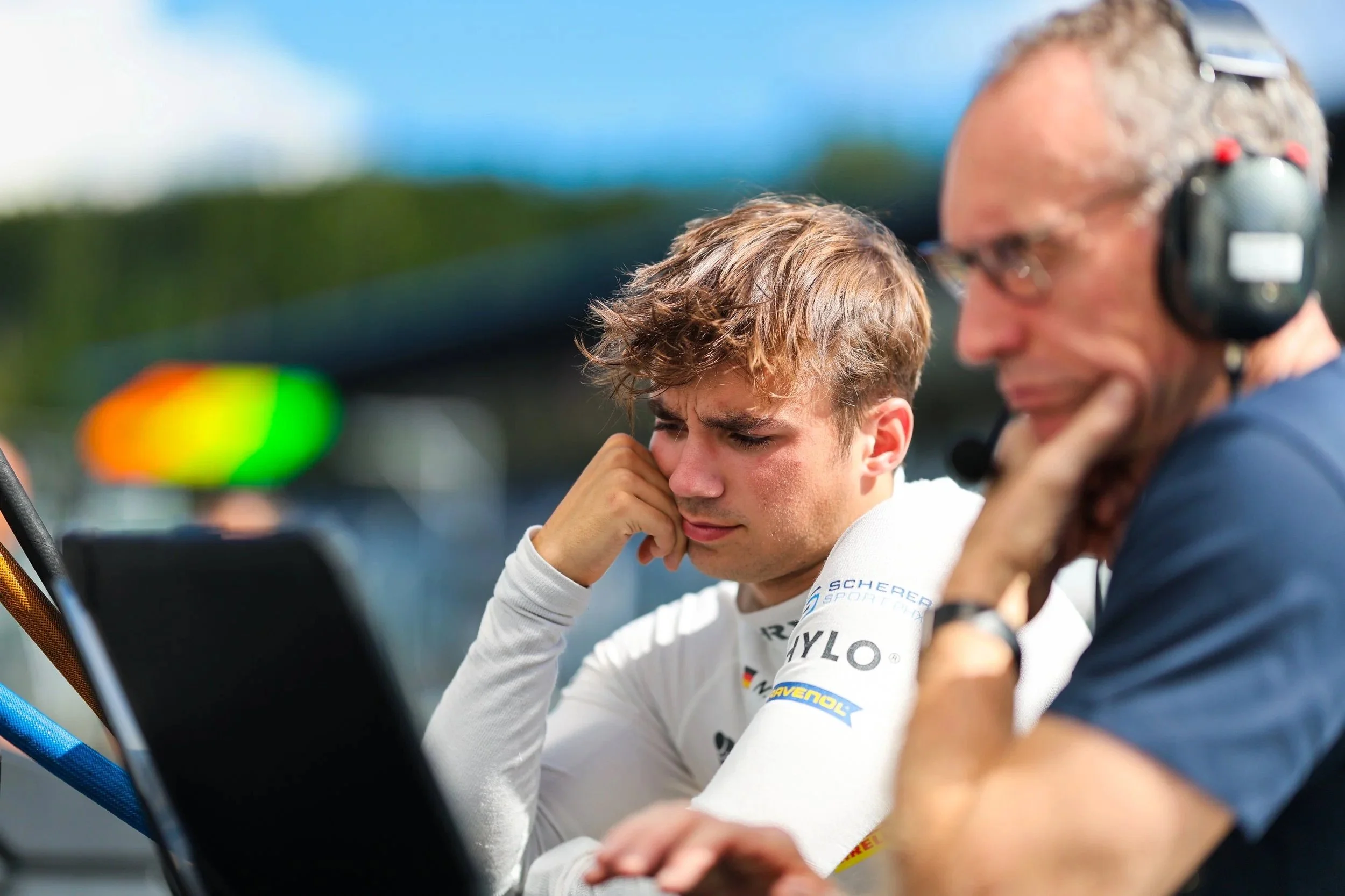 Ein junger Rennfahrer mit weißem Rennanzug und braunem, lockigem Haar sitzt nach einem Rennen mit Kopfhörer im Paddock, wirkt nachdenklich, während ein älterer Mann mit Brille und Headset neben ihm arbeitet.