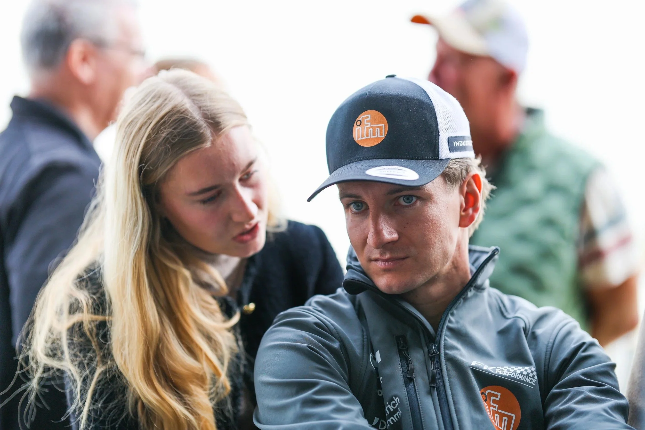 Junge Männer und Frauen im Gespräch, einer trägt eine schwarze Jacke und eine schwarze Cap mit Logo, andere haben blonde Haare und casual Kleidung, im Hintergrund weitere Personen, vermutlich bei einem Event oder Treffen.