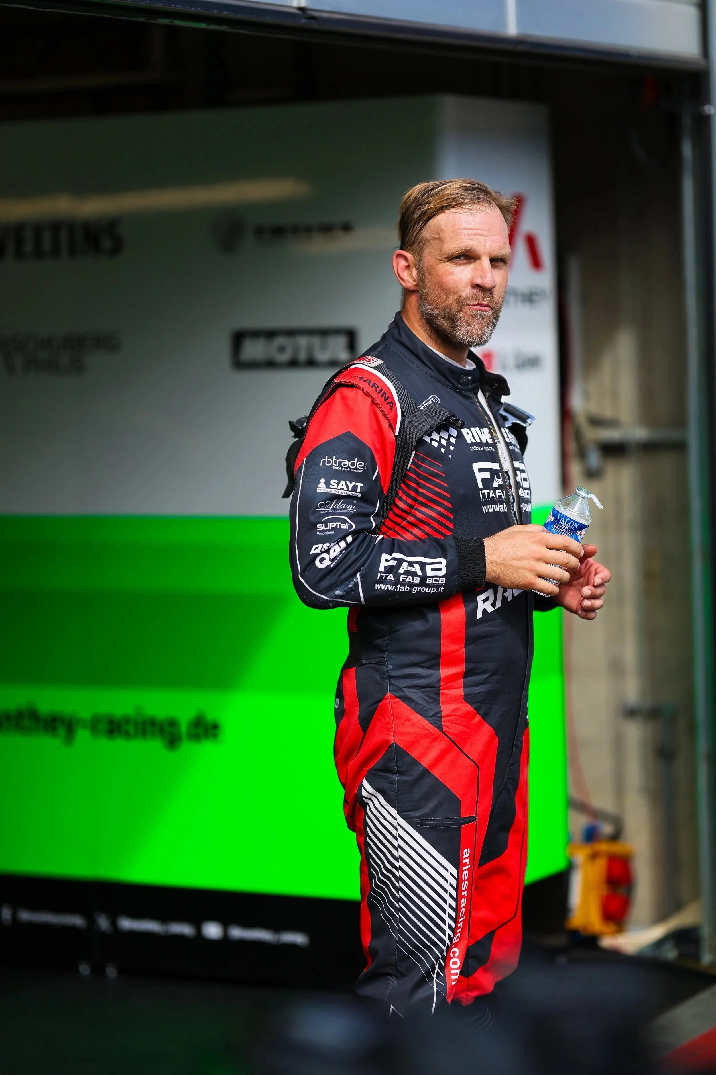 Ein Mann im roten und schwarzen Motorsport-Uniform steht mit einer Wasserflasche in der Hand und blickt nach vorne.