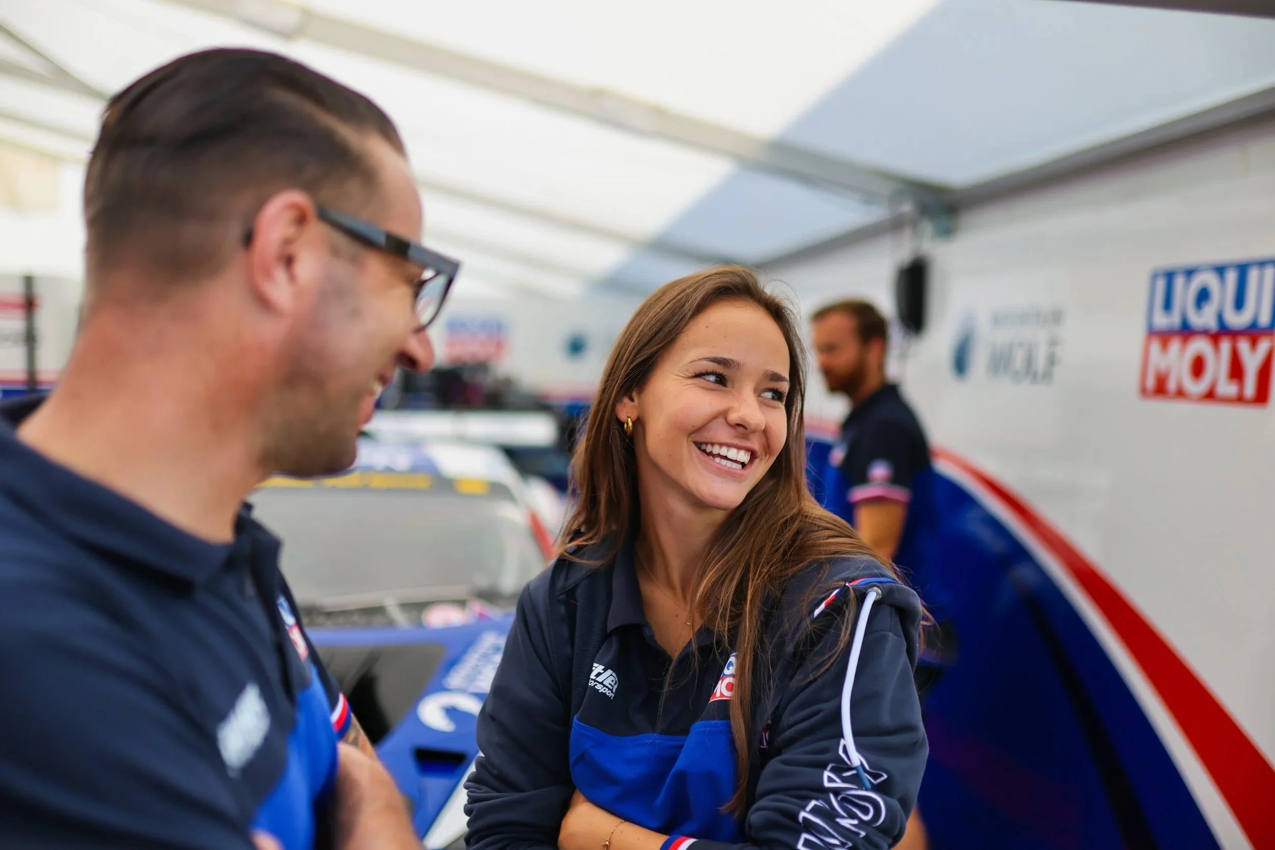 Motorsportteam in einer Garage, lachende Frau und lachender Mann in Teamkleidung mit Markenlogos, im Hintergrund weitere Teammitglieder und Rennwagen.