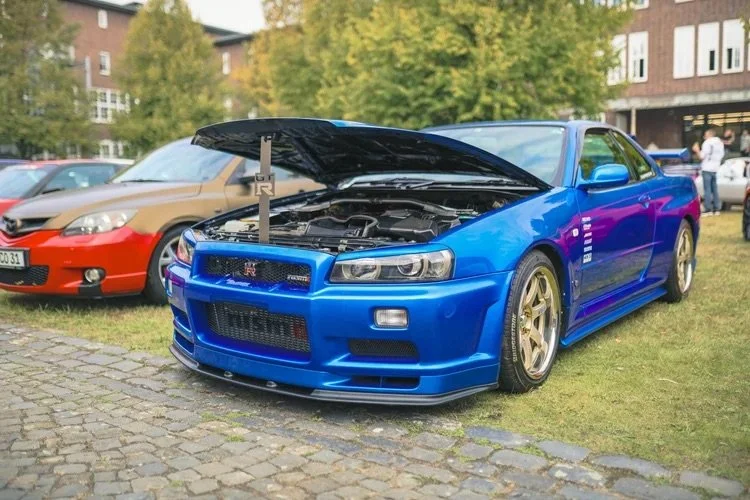 Blauer Nissan Skyline GT-R mit offenem Motorhaube auf Wiese bei Auto-Show, im Hintergrund weitere Fahrzeuge und Menschen