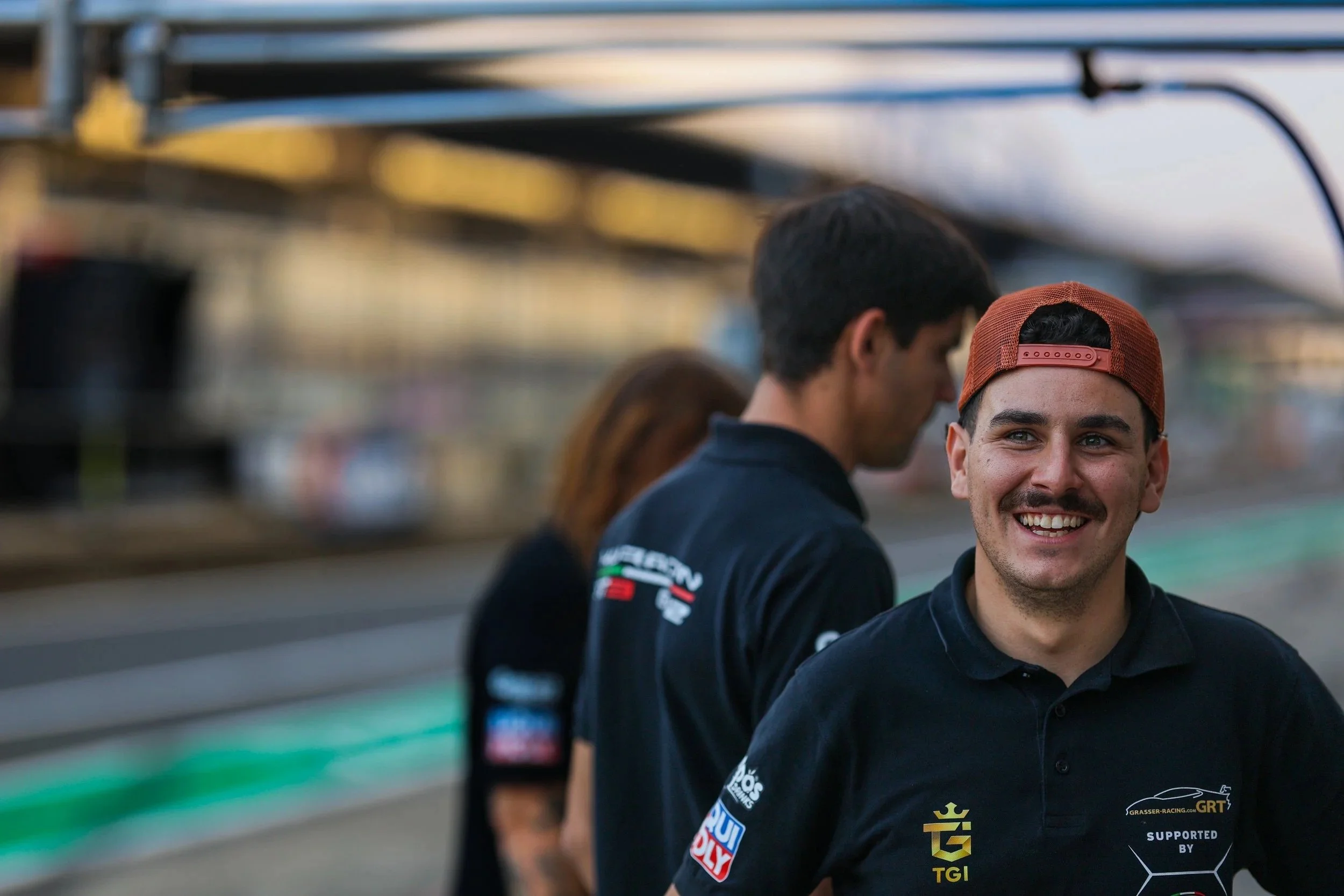 Junger Mann trägt schwarze Polo mit logos, rote Kappe rückwärts, lächelnd, im Motorsport-Umfeld, im Vordergrund; im Hintergrund zwei weitere Personen, unschärfer, ebenfalls im Motorsport-Design, an einer Rennstrecke.