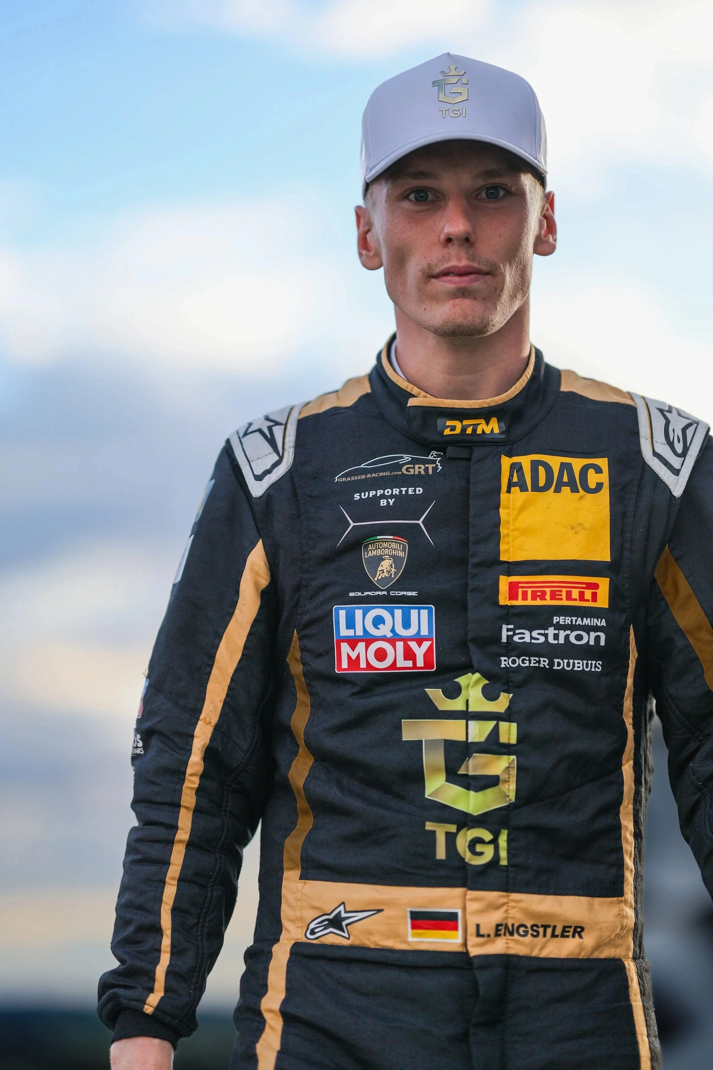 Junger Rennfahrer in schwarzem Motorsportanzug mit goldenen Akzenten und mehreren Sponsorenlogos, trägt graue Cap, steht draußen bei bewölktem Himmel.