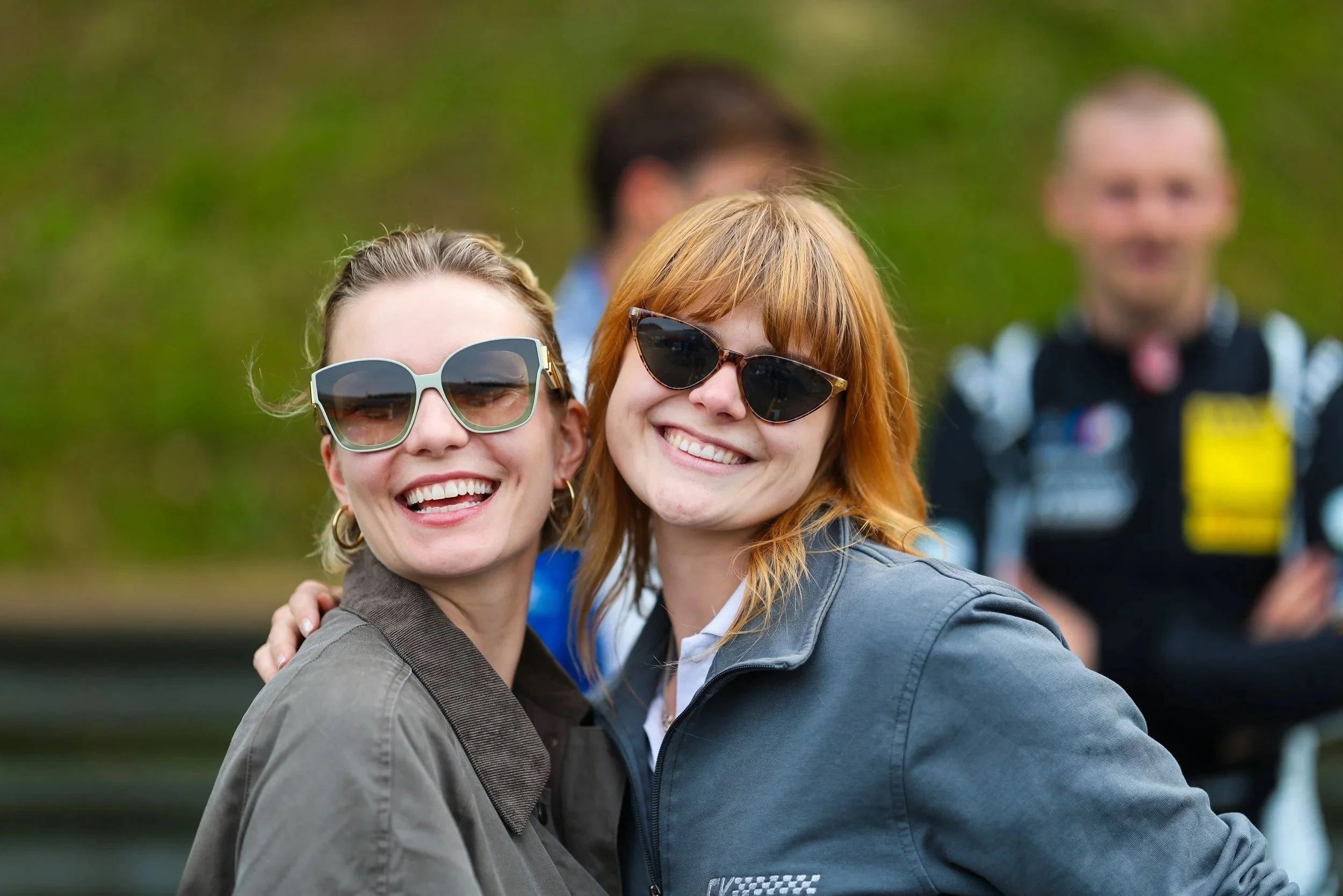 Zwei lachende Frauen mit Sonnenbrillen, die sich umarmen, im Freien bei einem Treffen mit Freunden.