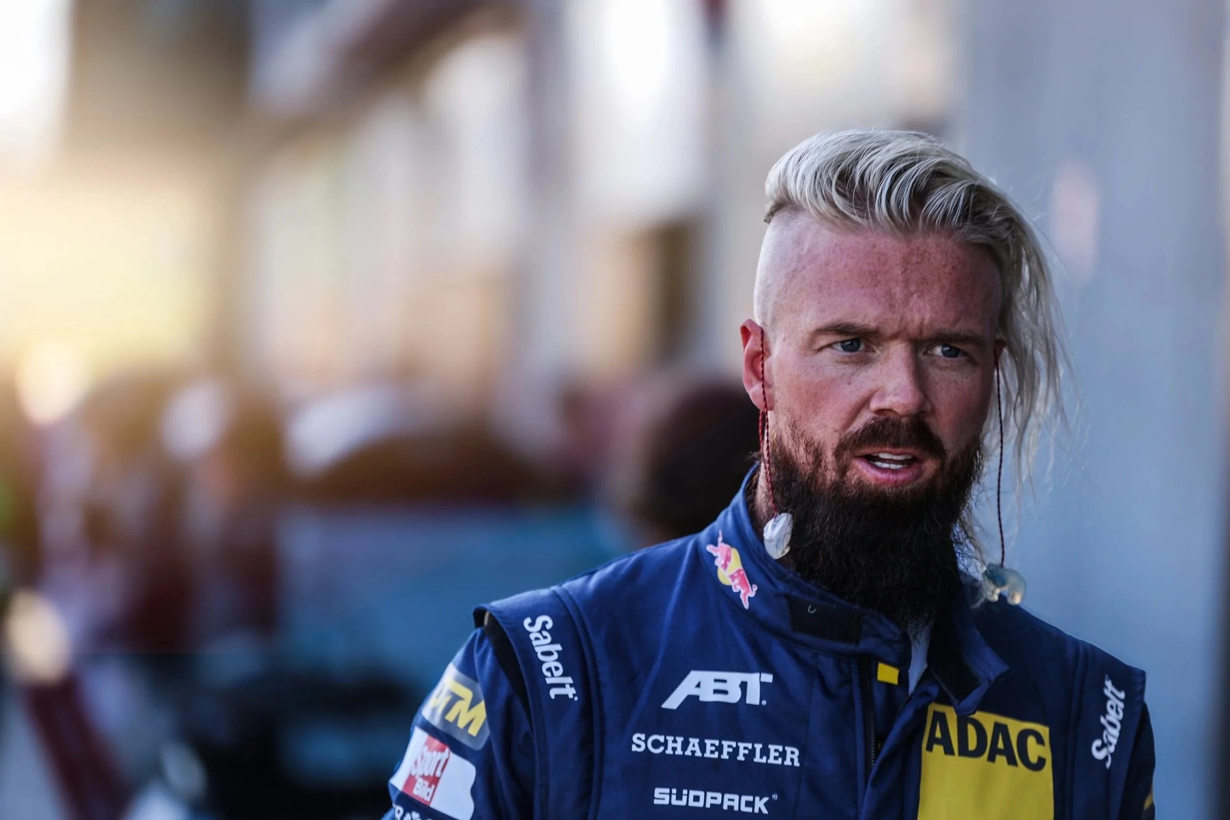 Ein Mann im Rennfahrer-Outfit mit blonder, seitlich gescheitelter Frisur, Bart und Earpods, stehend in einer Boxengasse oder bei einem Motorsport-Event.