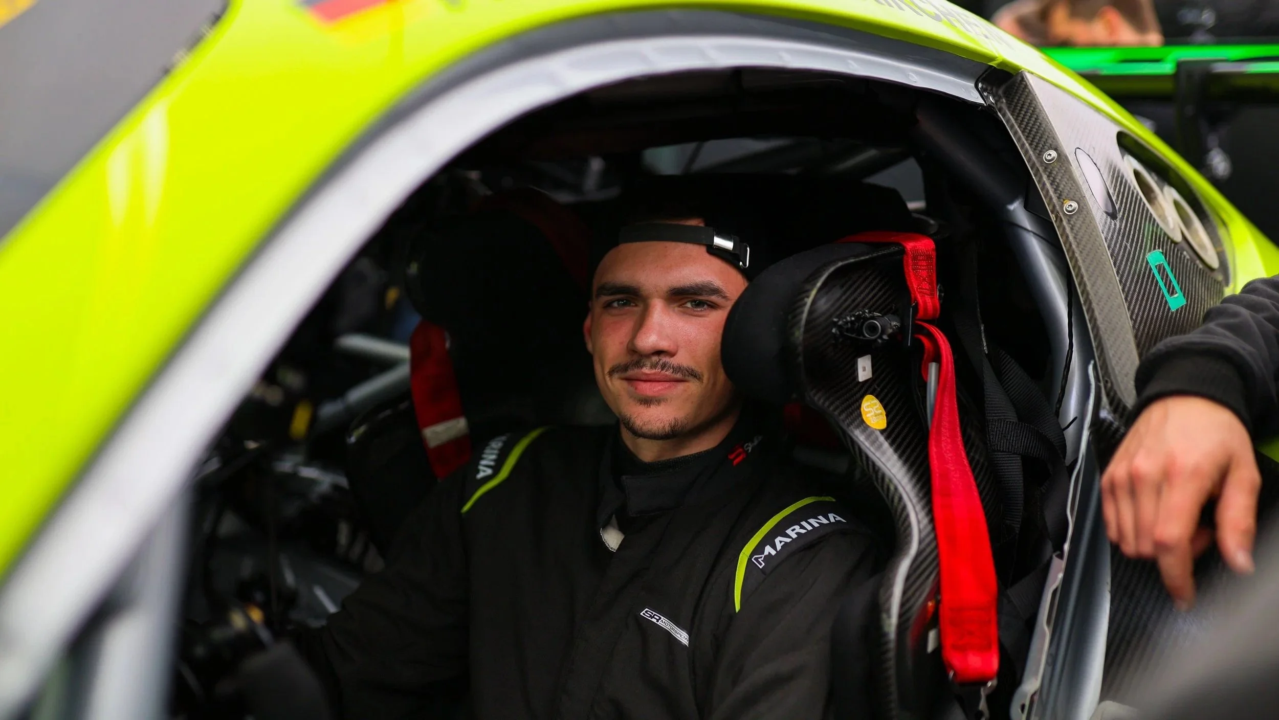Motorsportfahrer im Rennwagen, trägt schwarze Rennbekleidung und sitzt im Cockpit, schaut in die Kamera.