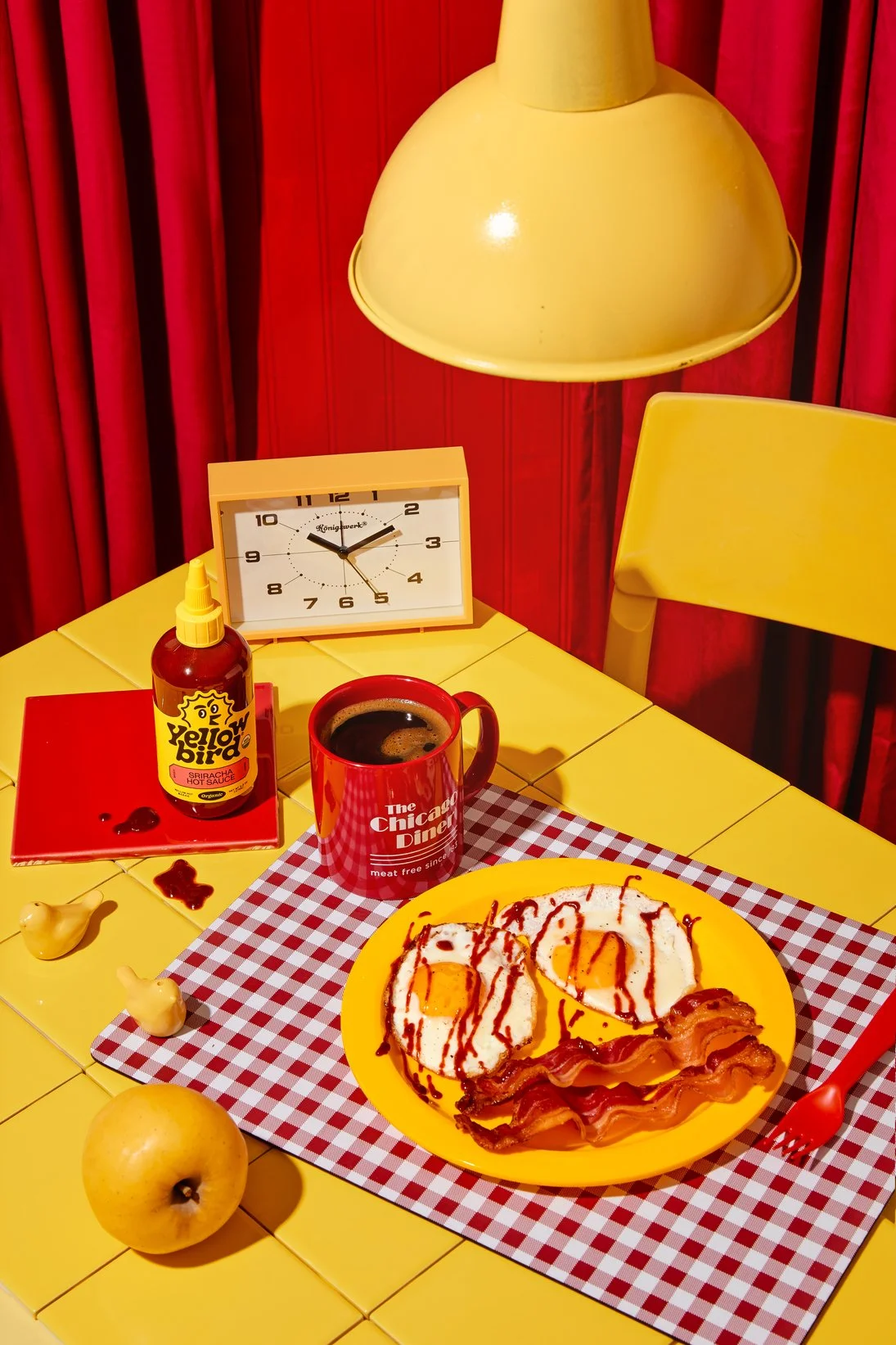 A breakfast setting with a yellow tiled table, yellow chair, yellow pendant light, red curtain, weather clock, red plate with eggs, bacon, and ketchup, a yellow apple, a red mug with coffee, a bottle of yellowbird hot sauce, a red fork, and two small