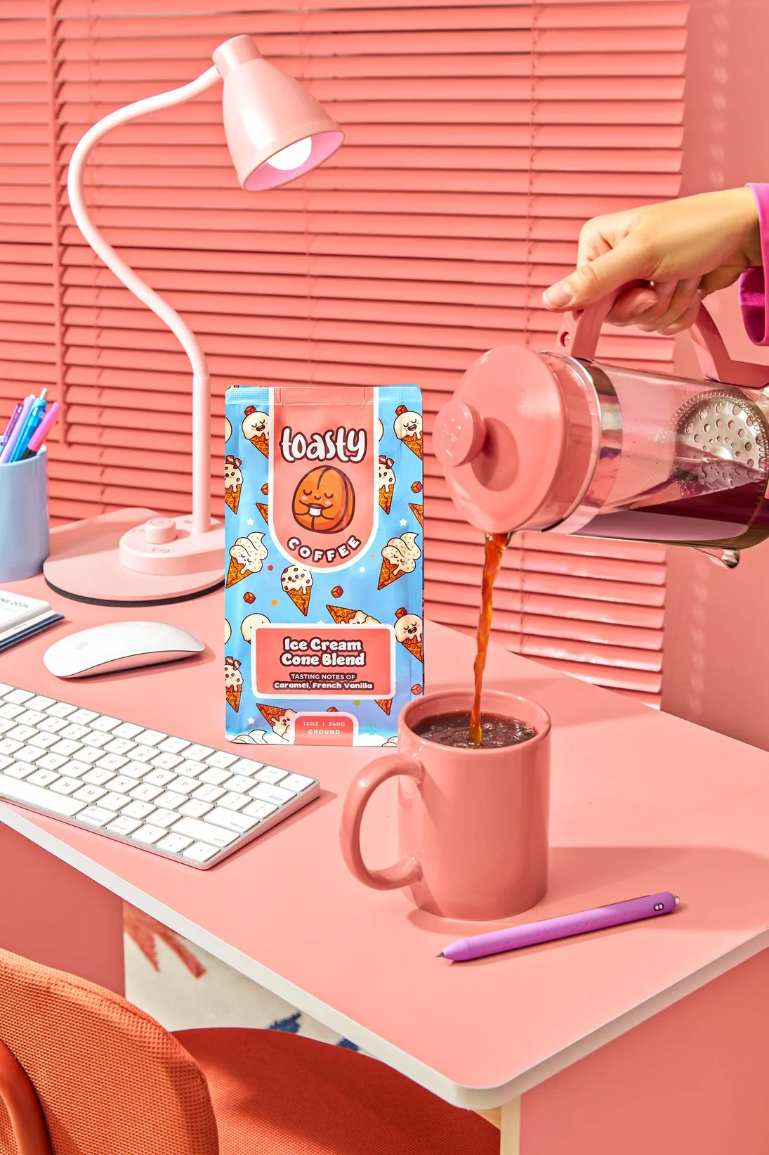 Pink desk setup with a pink lamp, white keyboard, mouse, and purple pen. A pink mug is being filled with coffee from a glass pot. Behind it is a carton of 'Tasty Coffee' ice cream cone blend, featuring cartoon ice cream cones and an orange character 