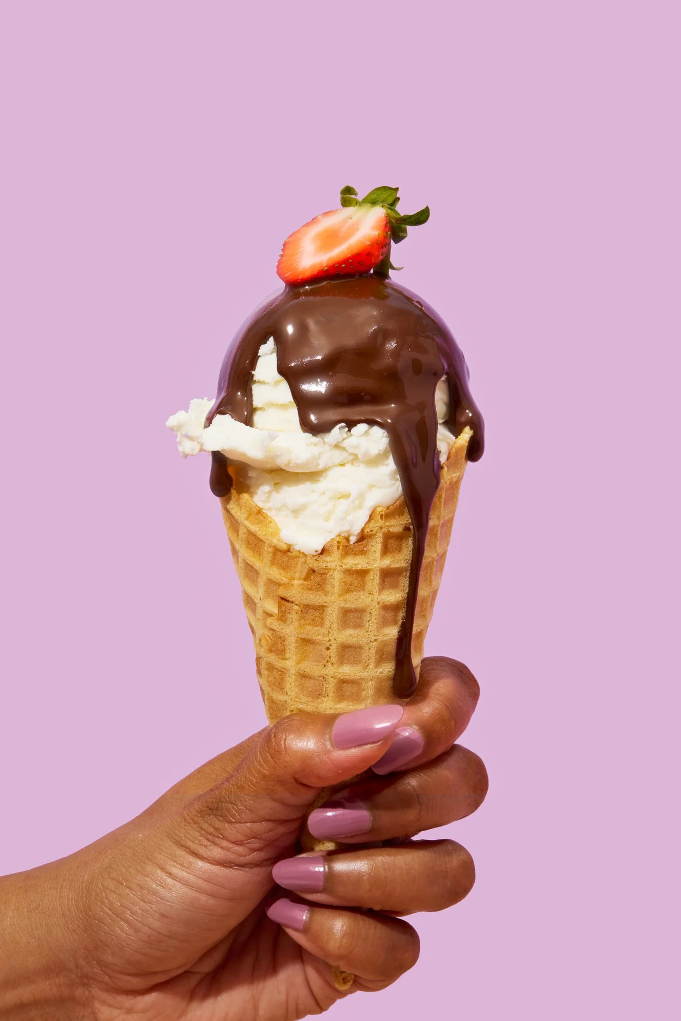 Hand holding an ice cream cone topped with strawberries and chocolate syrup against light purple background.