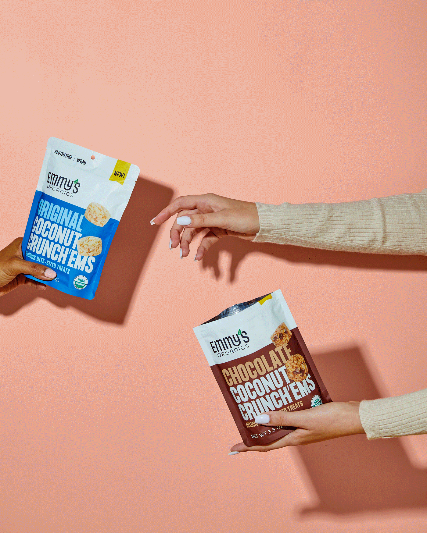 Two hands holding packages of coconut crunch treats, one with blue and white packaging labeled 'Original Coconut Crunch,' the other with brown and white packaging labeled 'Chocolate Coconut Crunch,' against a pink background.