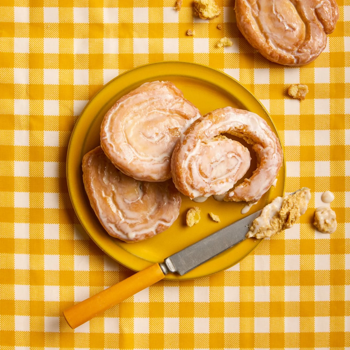Danish pastries with icing on a yellow plate, placed on a yellow and white checkered tablecloth, with scattered crumbs and a butter knife.