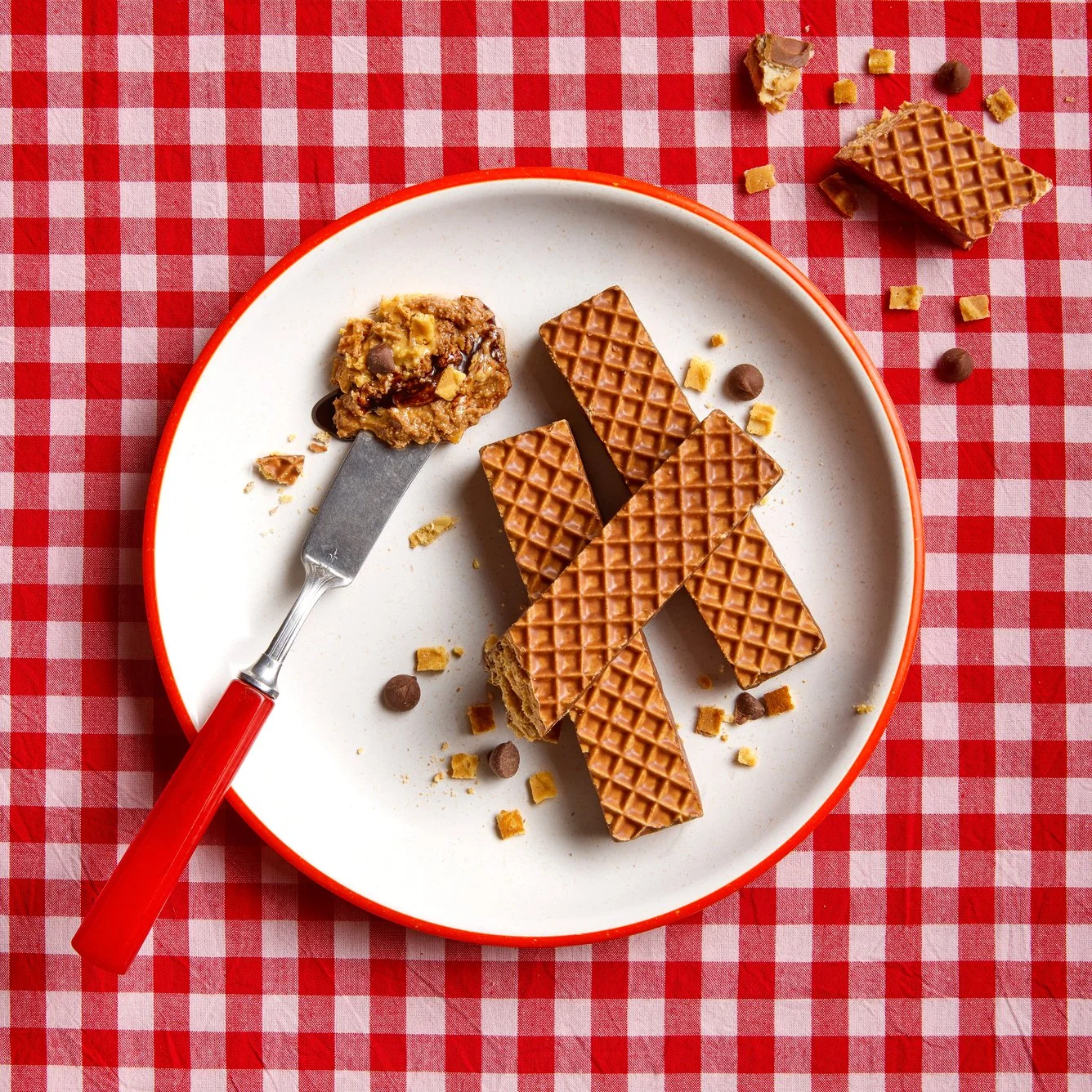 A white plate with a red rim sitting on a red and white checkered tablecloth, containing broken pieces of caramel-flavored wafer bars, a cookie with chocolate chips, and scattered chocolate chips and cookie crumbs.