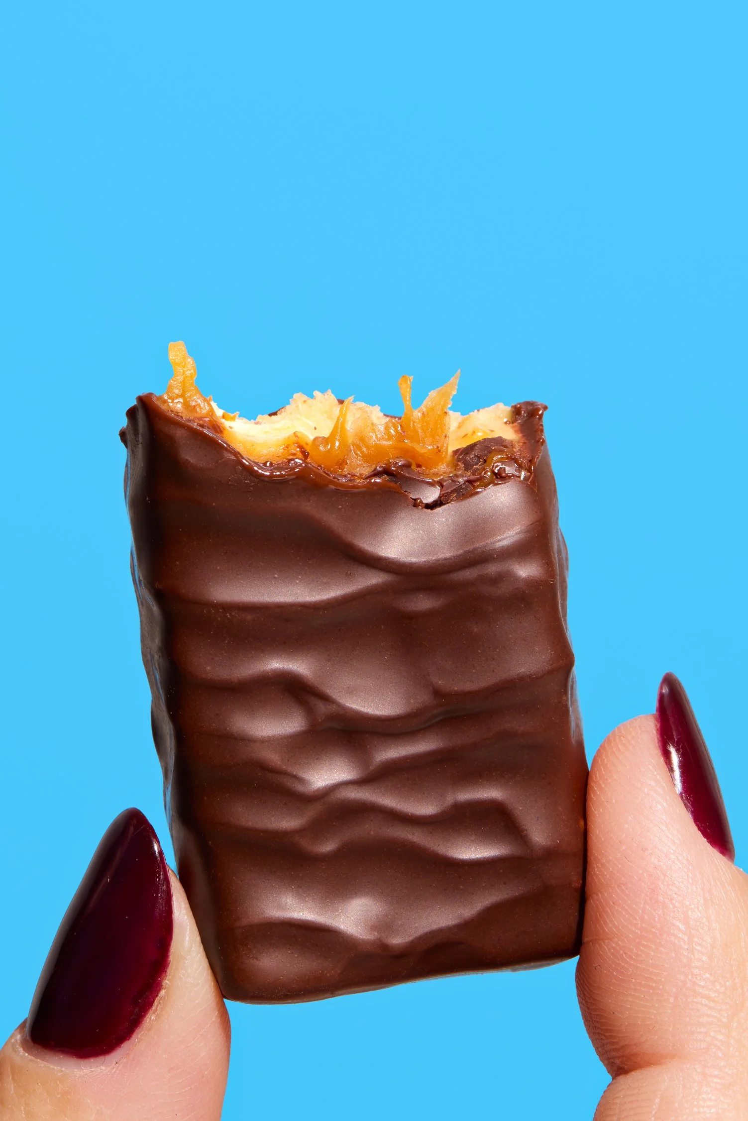 Close-up of a chocolate-covered candy bar with caramel and nuts, held between a person's thumb and index finger against a blue background.