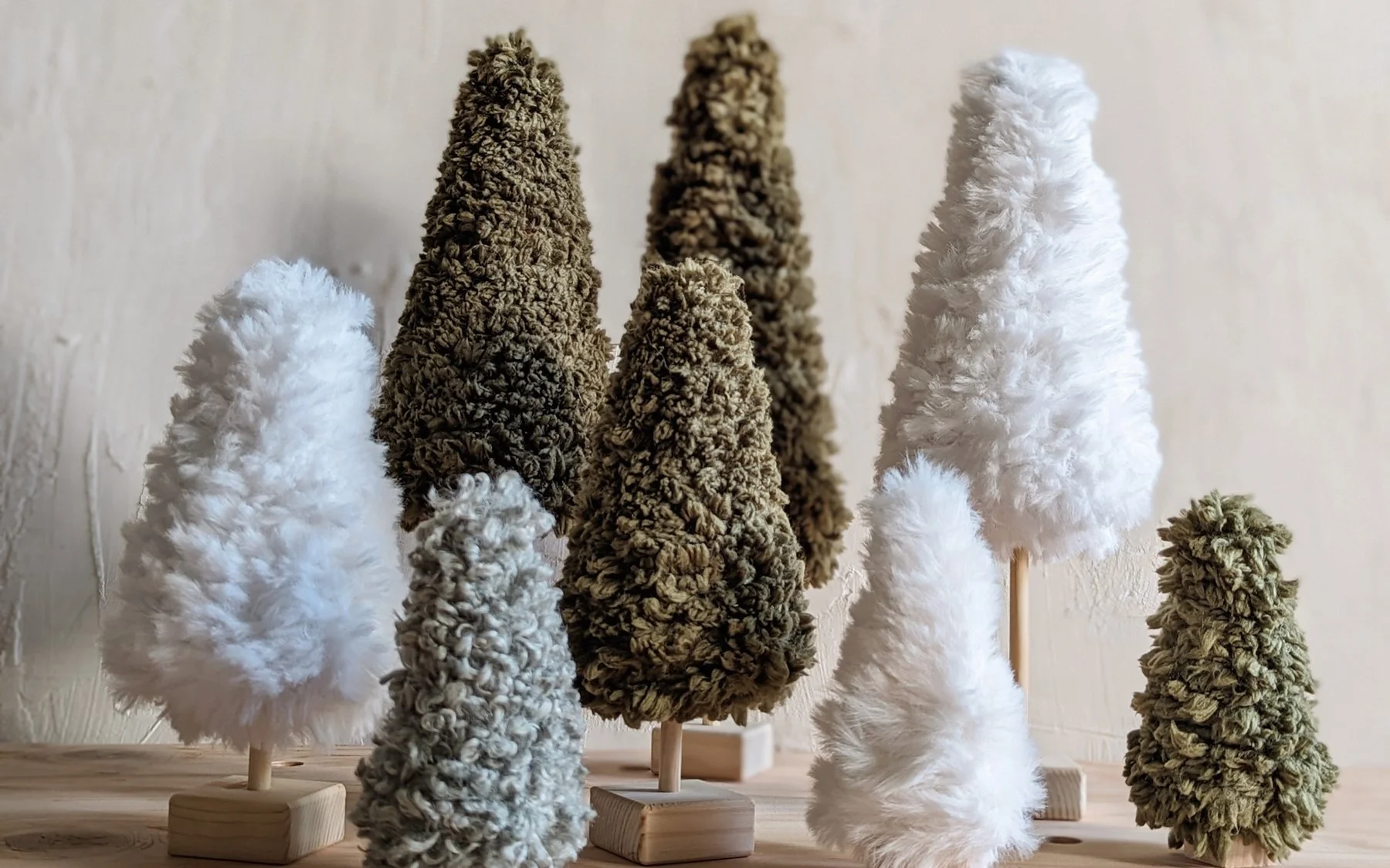 yarn-y trees in green, sage, and white furn yarn stand in a line in front of a textured background