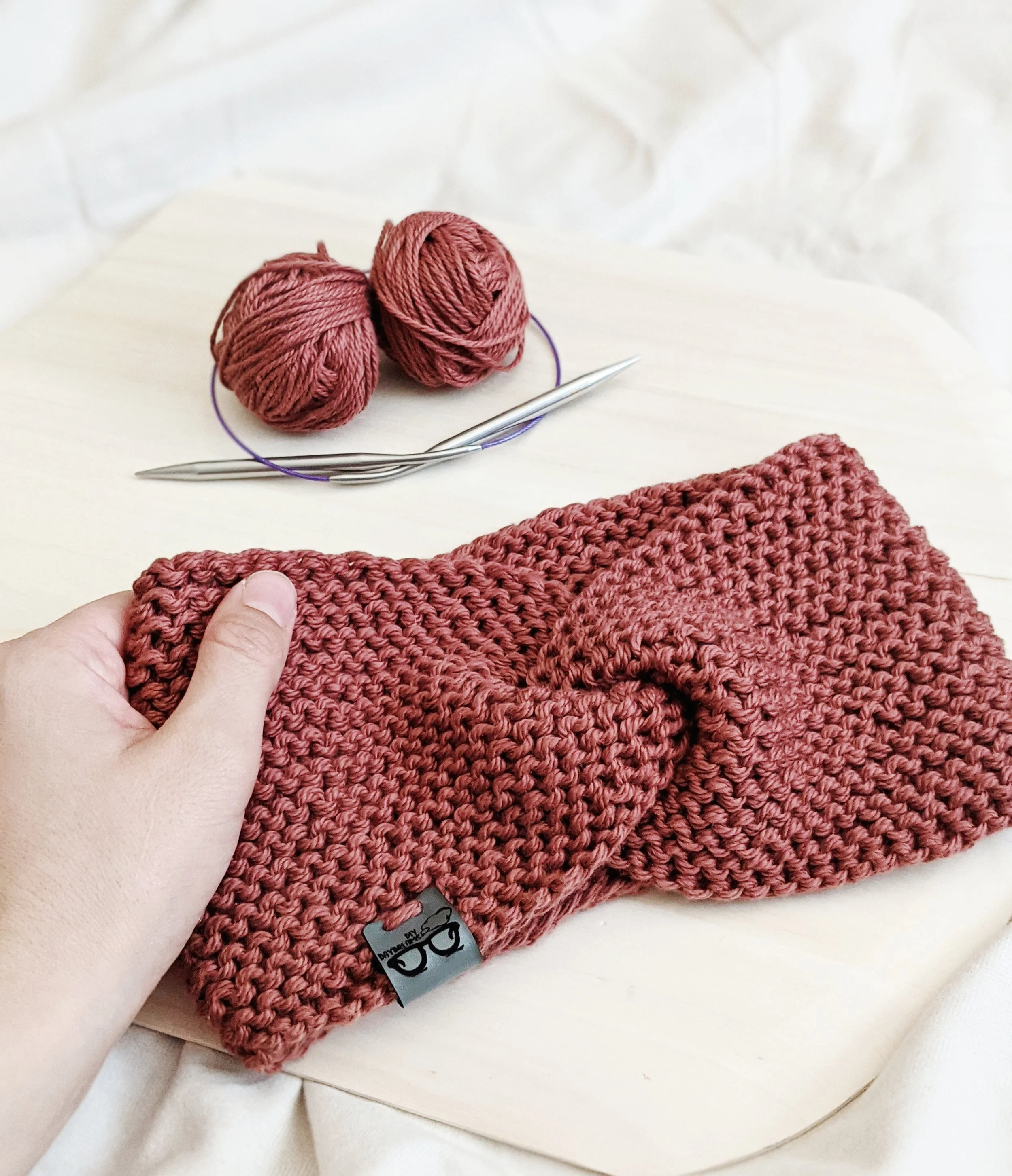 a cotton deluxe twist headband is laying on a piece of wood with a hand gently holding it while knitting needles and some leftover yarn sit nearby