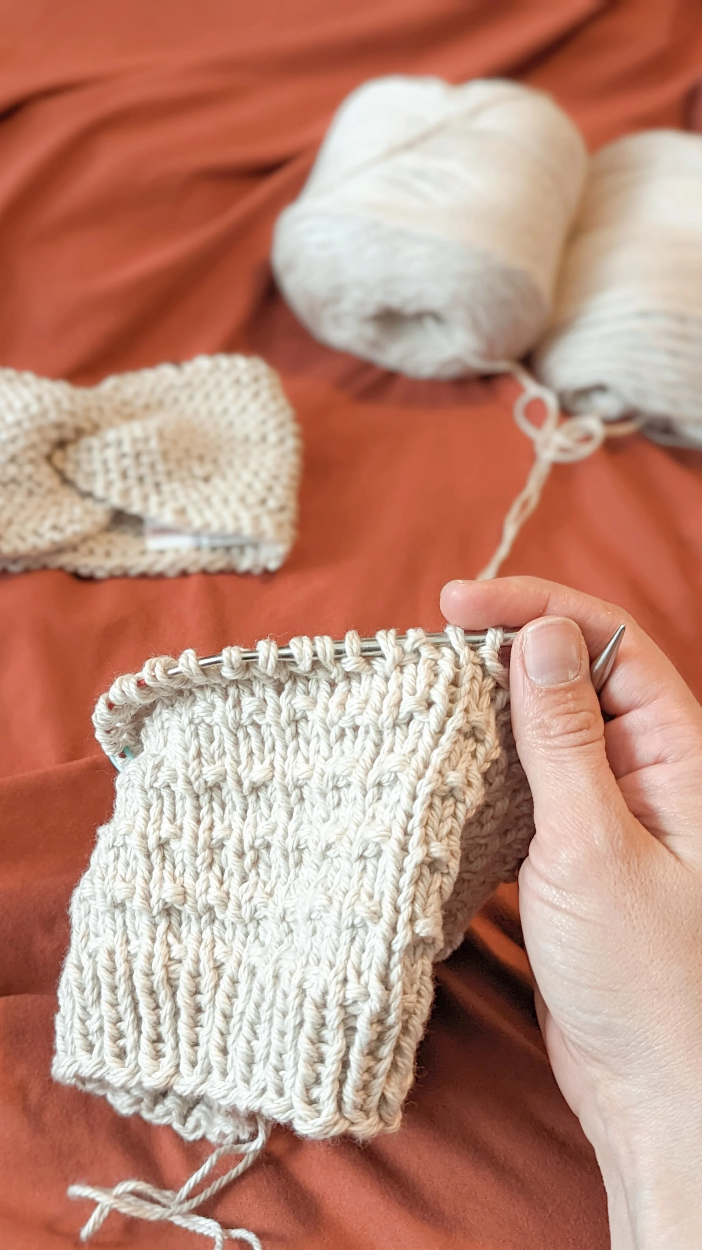 a linen cotton waffle beanie is half knit on a rust-colored background. a hand holds the knitting needles and balls of yarn are seen in the background
