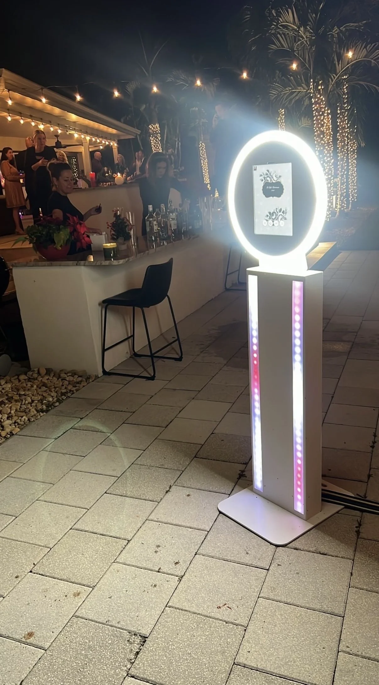 An outdoor bar at night with a group of people in the background. A circular illuminated photo booth stands on the paved ground in the foreground, emitting bright white and pinkish-purple lights.