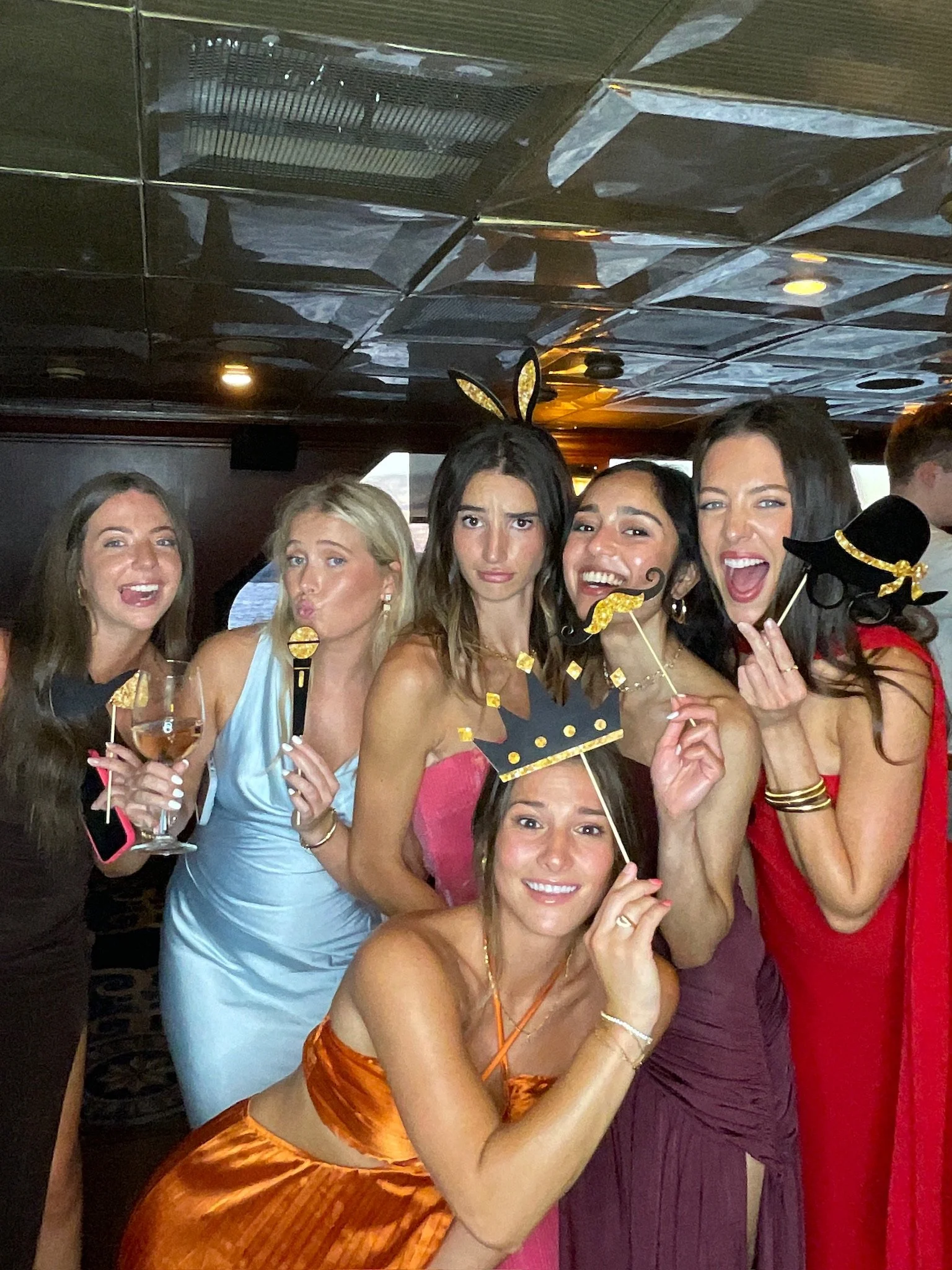 A group of women celebrating at a party, wearing colorful dresses, holding drinks, and using fun photo props like a crown, mustache, and hat. They are smiling and laughing.