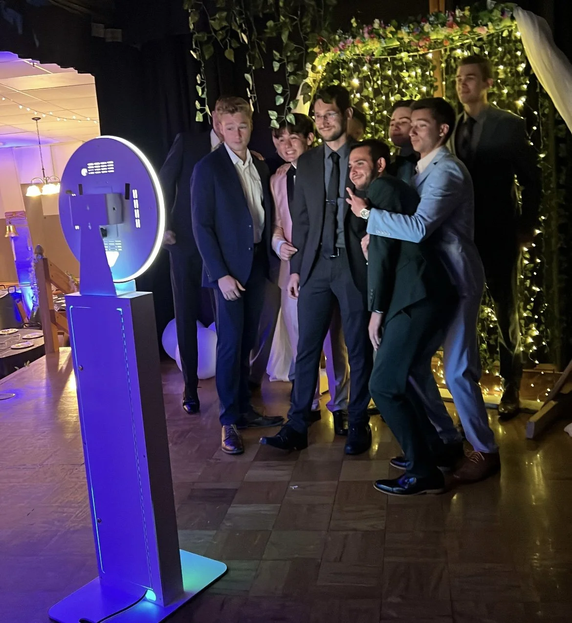 Group of men in formal suits taking a photo in front of a decorated backdrop with fairy lights and greenery, lit with colorful and white lights, in an indoor venue.